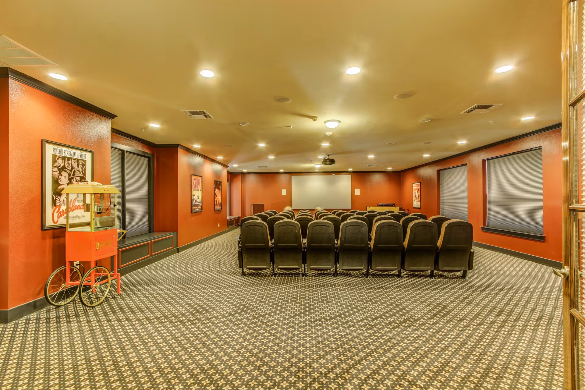 A senior living facility movie theater room with rows of black cushioned chairs facing a large white projection screen. The walls are painted red with black trim, decorated with vintage movie posters. There is a popcorn machine on the left side near the entrance, and the floor is covered with a patterned carpet.