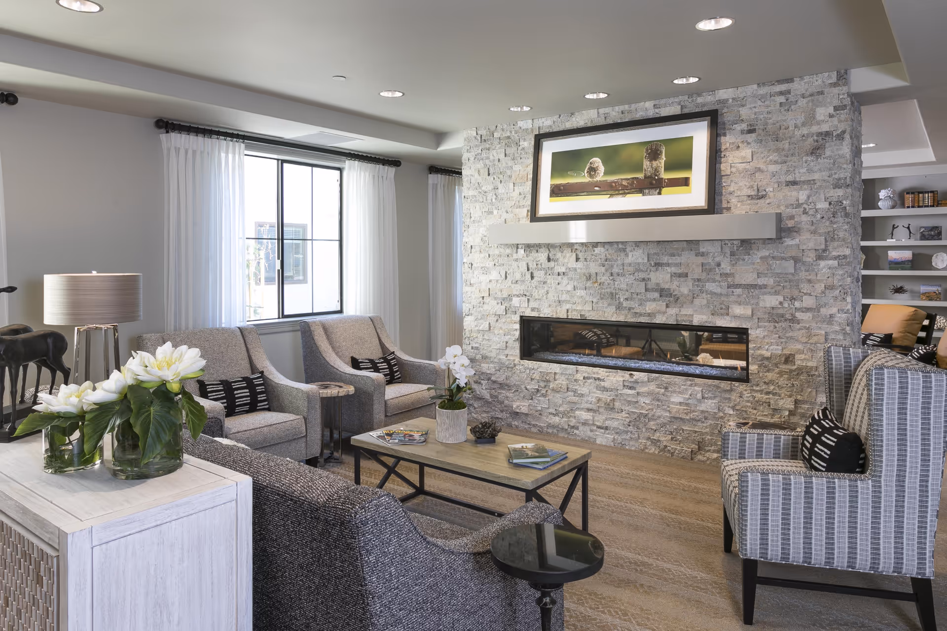 A modern living room with a stone accent wall featuring a long rectangular fireplace and a framed picture above it. The room has a large window with white curtains, two gray armchairs with black and white pillows, a gray sofa, a wooden coffee table with books and a plant, and a side table with a lamp and white flowers in glass vases.