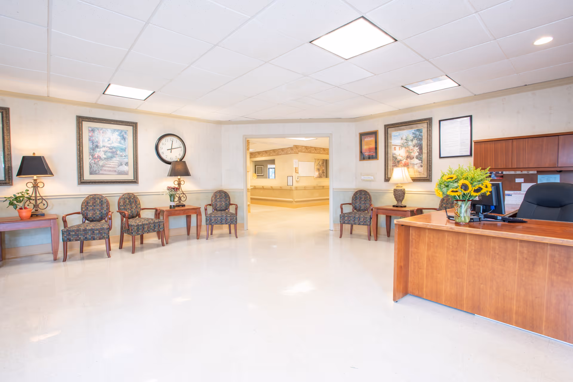 A bright and clean reception area in a healthcare facility with a wooden desk on the right adorned with a vase of sunflowers. Several patterned chairs are arranged along the walls with small tables holding lamps and plants. Framed artwork and a clock decorate the walls. An open doorway leads to a hallway with handrails.