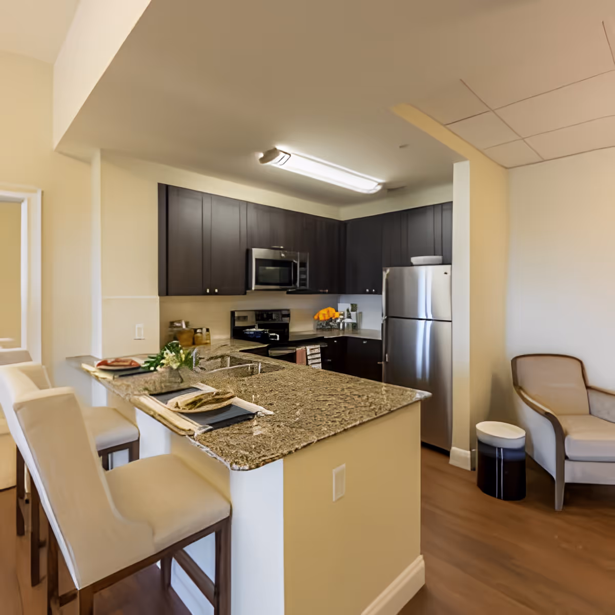 Modern kitchen area with dark wood cabinets, stainless steel refrigerator and microwave, granite countertop with two place settings, and two beige upholstered bar stools. Adjacent to the kitchen is a beige armchair and a small round side table.