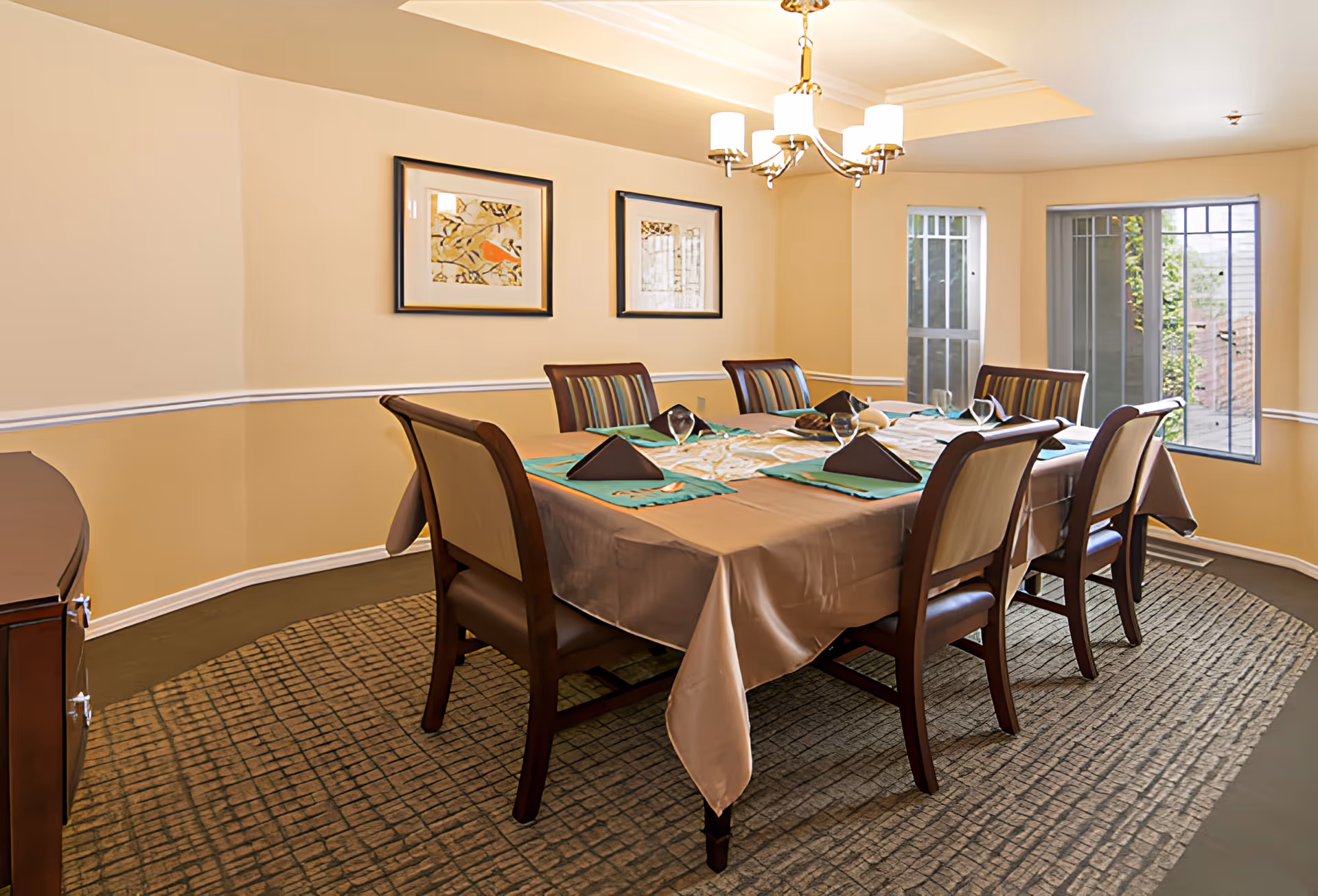 A dining room with a rectangular table covered with a brown tablecloth and set with green placemats, folded black napkins, and glassware. Six wooden chairs with cushioned seats surround the table. The room has beige walls with white trim, two framed pictures on the wall, a chandelier hanging from the ceiling, and two windows letting in natural light.