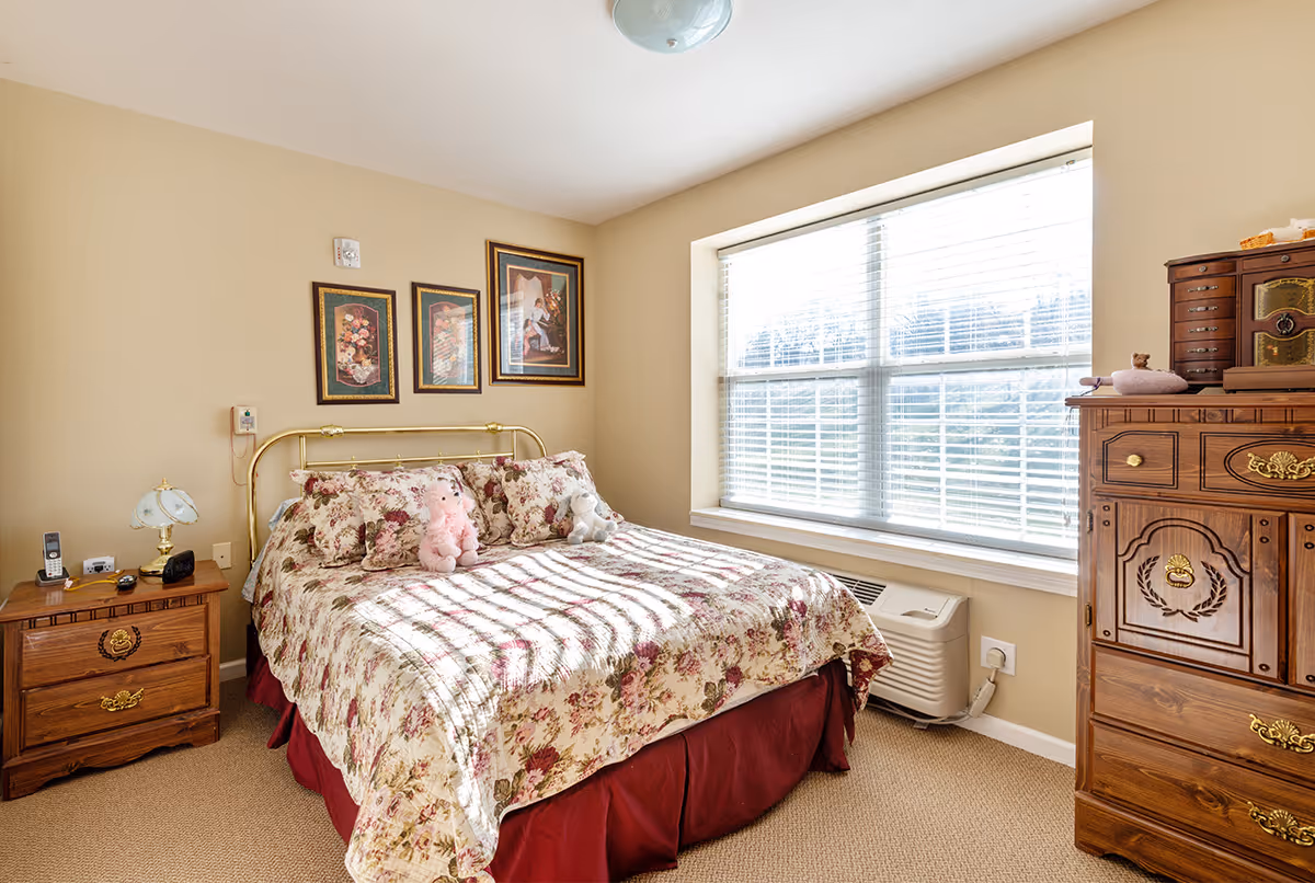 A cozy bedroom with a floral bedspread on a double bed, two stuffed animals on the bed, a wooden nightstand with a lamp and phone, three framed pictures on the wall, a large window with blinds letting in sunlight, and a wooden dresser with decorative items on top.