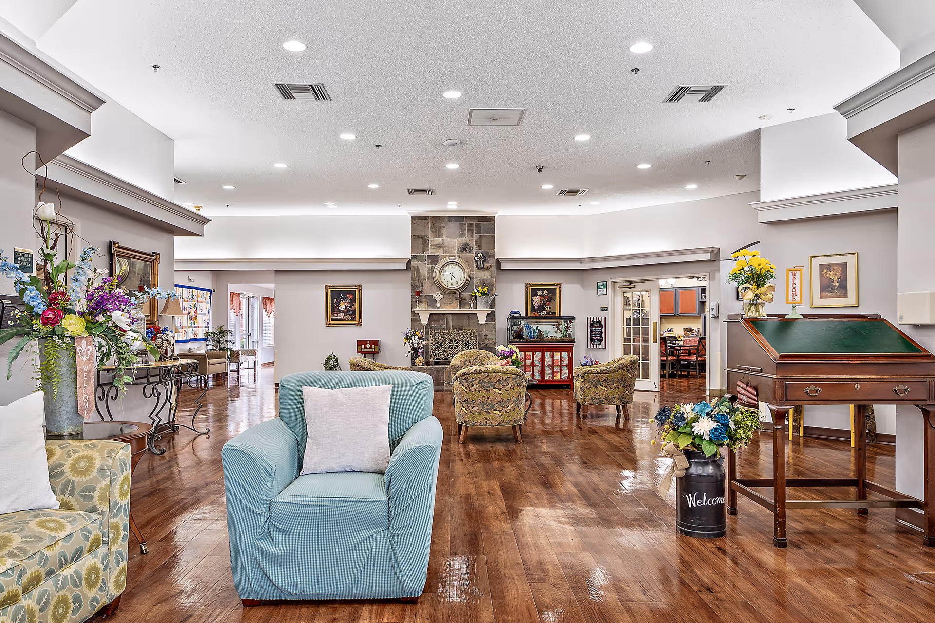 A spacious senior living facility common area with wooden floors, a stone fireplace with a clock above it, and several upholstered chairs arranged around the room. There are floral arrangements on tables and a wooden writing desk on the right side. The room is well-lit with recessed ceiling lights and decorated with framed artwork on the walls.