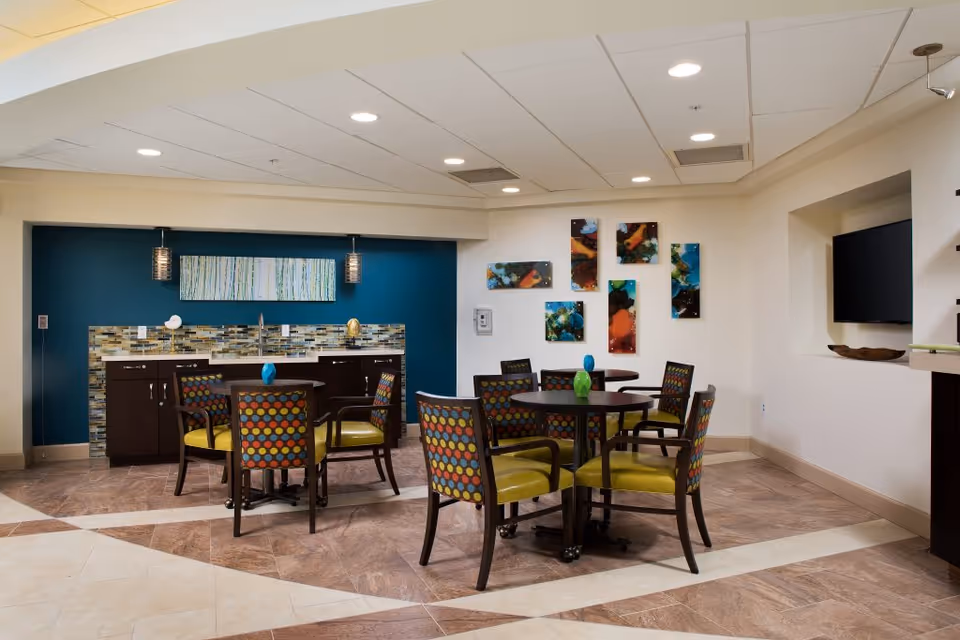 A modern common area with round tables and colorful chairs featuring green seats and multicolored patterned backs. The back wall has a blue accent with a mosaic tile backsplash and dark wood cabinets. Above the cabinets is a horizontal abstract painting. On the adjacent wall, there are several abstract art pieces arranged in a grid. The ceiling has recessed lighting, and a flat-screen TV is mounted in a recessed wall niche.