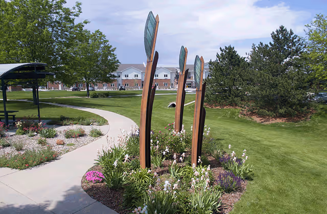 Landscaped courtyard with a curved concrete path, tall metal sculptures, flowerbeds, and a multi-story facility building in the background.