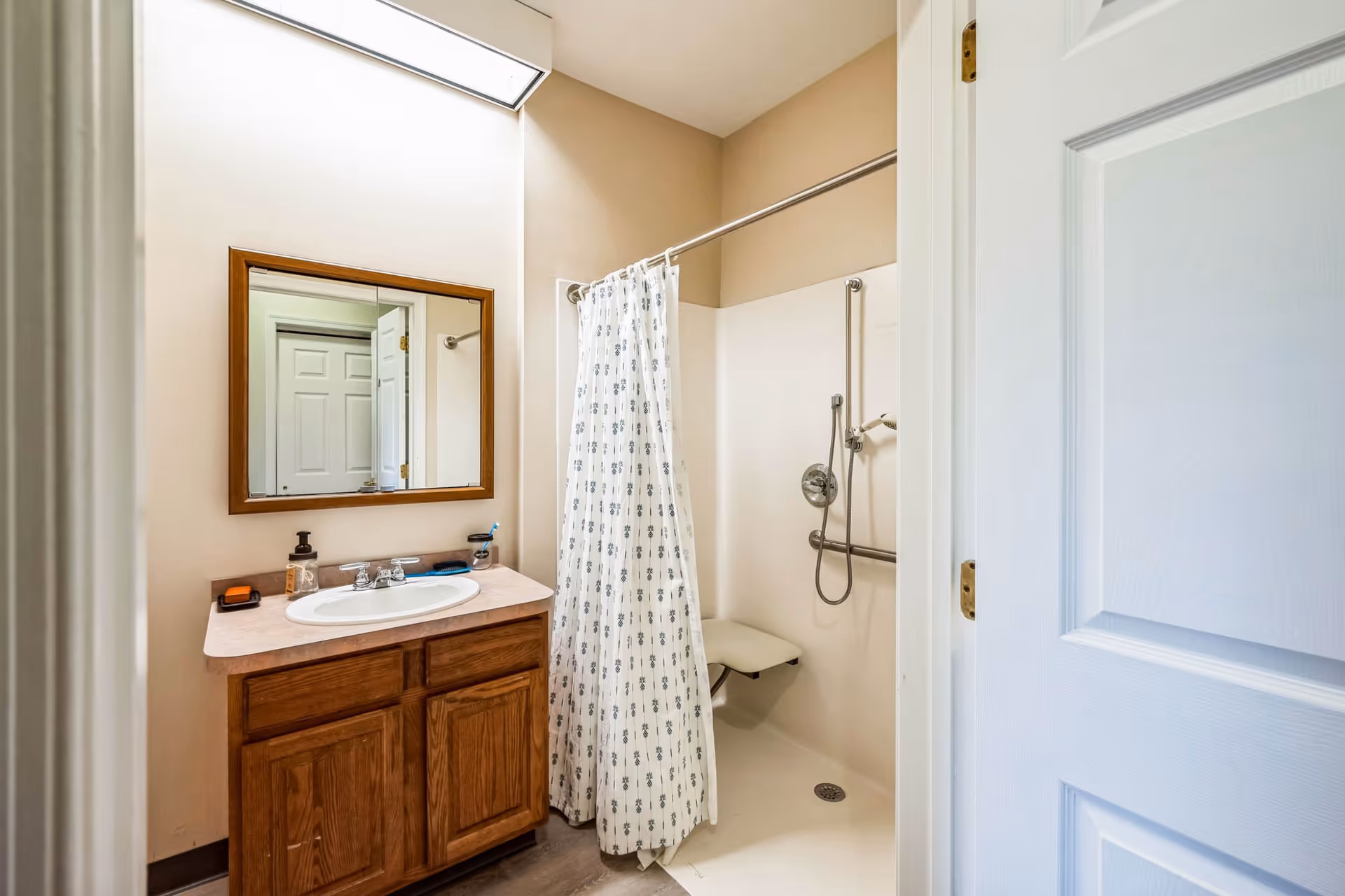 A bathroom with a wooden vanity cabinet and a sink topped with a mirror. Next to the vanity is a walk-in shower with a white curtain featuring a subtle pattern, a fold-down shower seat, and grab bars for accessibility. The room has beige walls and a white door partially open.