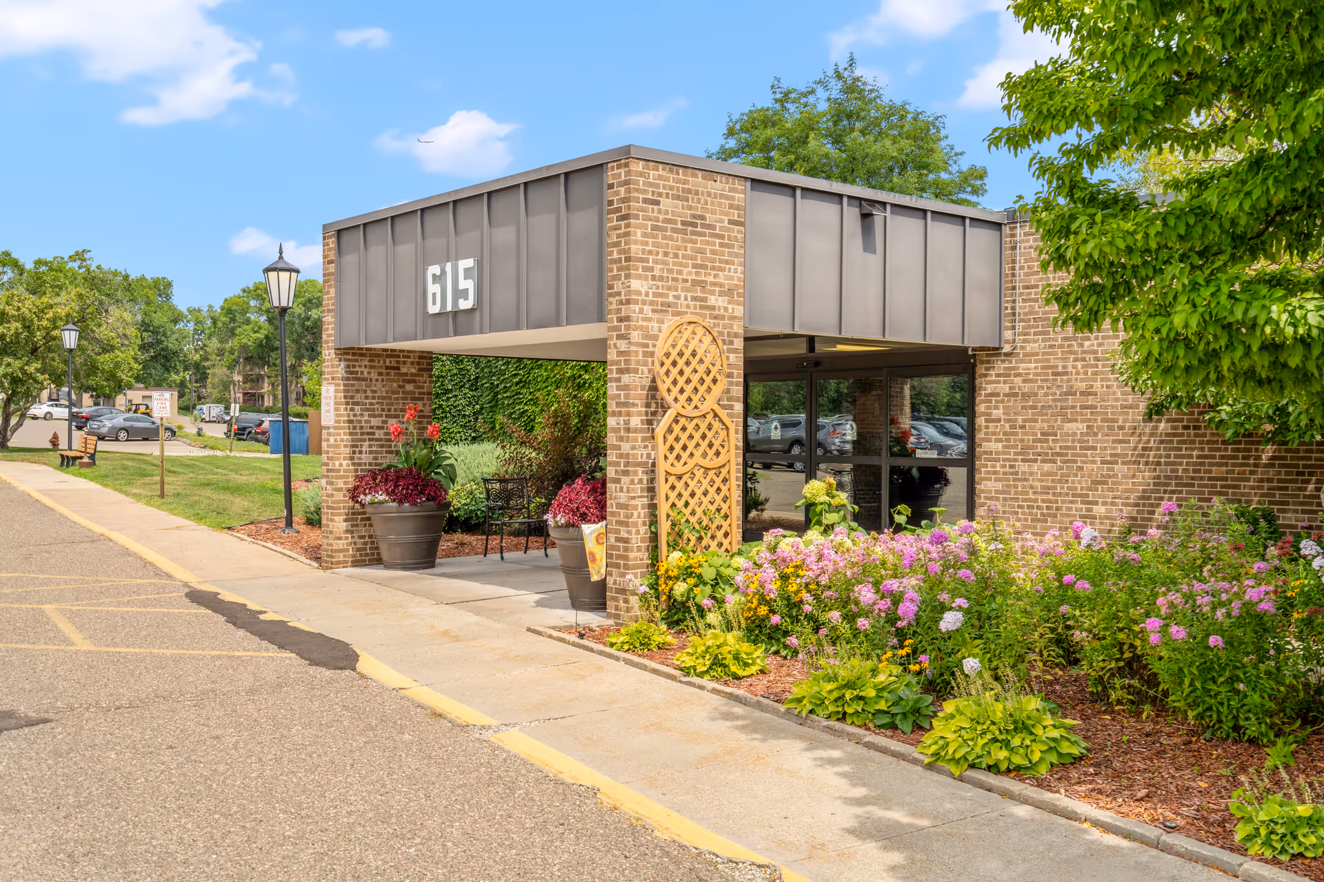 Entrance of a brick building with the number 615 displayed above a covered walkway. There are flower beds with colorful flowers and greenery along the sidewalk, and a bench and lamp posts are visible in the background under a blue sky with some clouds.