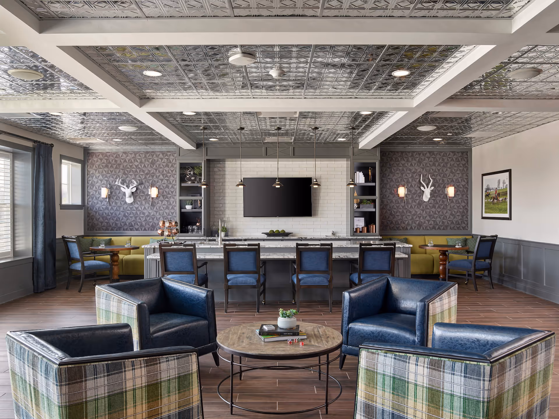 A modern lounge area with four blue leather and plaid armchairs arranged around a round wooden coffee table with books and a small plant. Behind the seating area is a long counter with six blue chairs, pendant lights hanging above, and a large flat-screen TV mounted on a white tiled wall. The room features decorative wall panels with white deer head sculptures and sconces, wood flooring, and a metallic patterned ceiling.