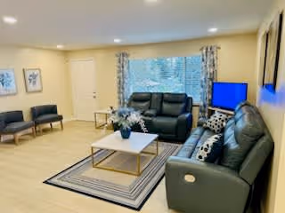 Well-lit living room with leather sofas, chairs, a coffee table on a rug, and a TV by a large window.