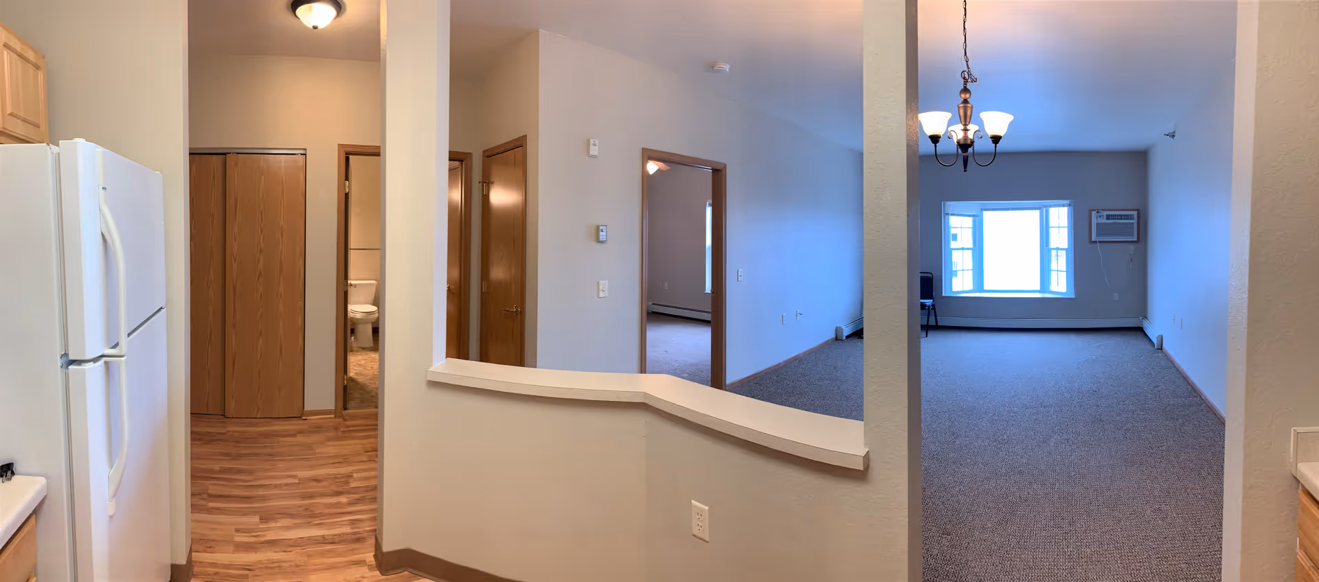 Open-plan interior showing a kitchen with refrigerator, a hallway with doors and a bathroom, and a carpeted living room with a window and chandelier.