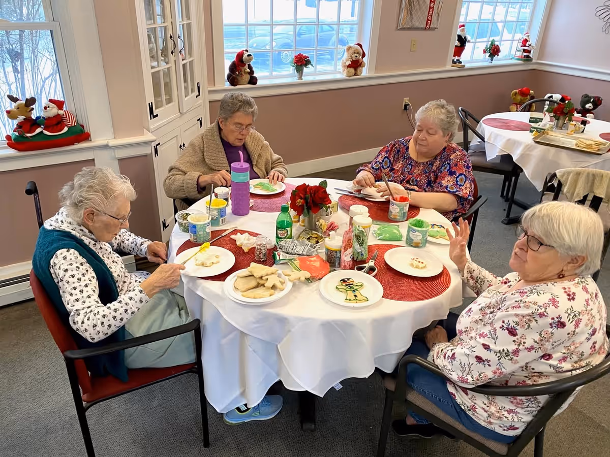 Four elderly women sitting around a round table in a dining area, decorating cookies with various colored icings and sprinkles. The table is covered with a white tablecloth and red placemats, and there are holiday decorations on the windowsills behind them.