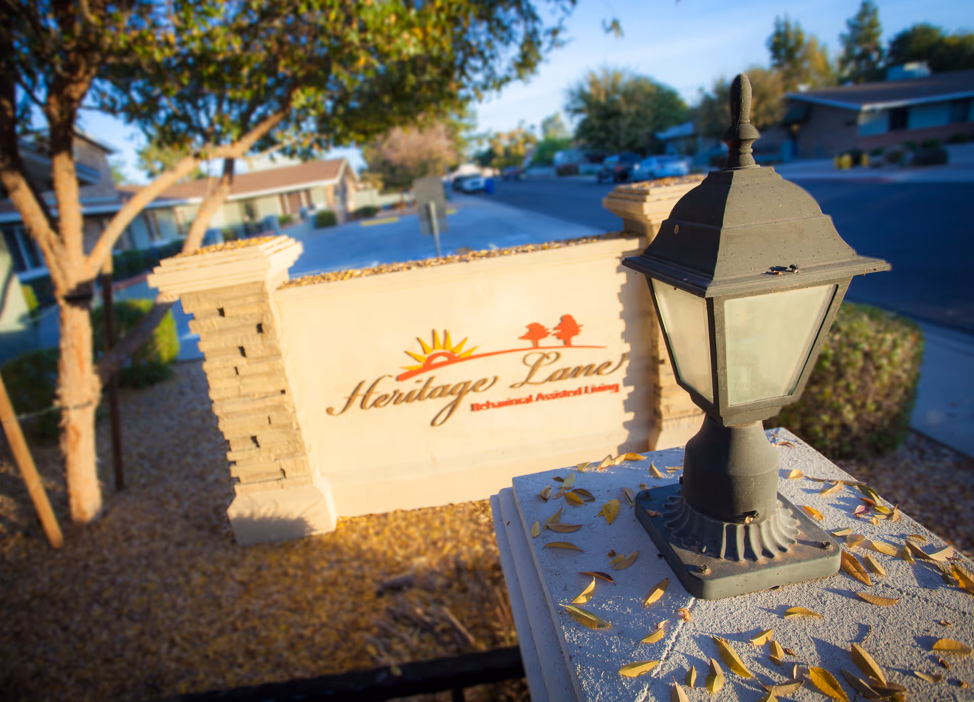 Outdoor view of the entrance sign for Heritage Lane Behavioral Assisted Living, featuring a stone sign with the facility's name and a decorative lamp post in the foreground, surrounded by trees and residential buildings.