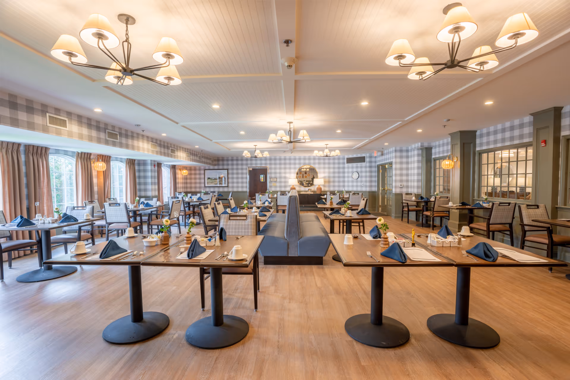 A spacious and well-lit dining room with multiple tables set for meals. The tables have blue napkins, white cups, and small flower arrangements. The room features large windows with curtains, patterned wallpaper, and modern chandeliers hanging from the ceiling. There is a central seating area with cushioned benches and chairs arranged neatly around the tables.