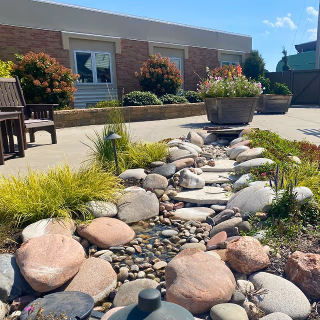 Outdoor garden area with a small decorative rock-lined water feature surrounded by plants and flowers. There are wooden chairs on a concrete patio next to a brick building with windows. Large planters with blooming flowers are also visible under a clear blue sky.