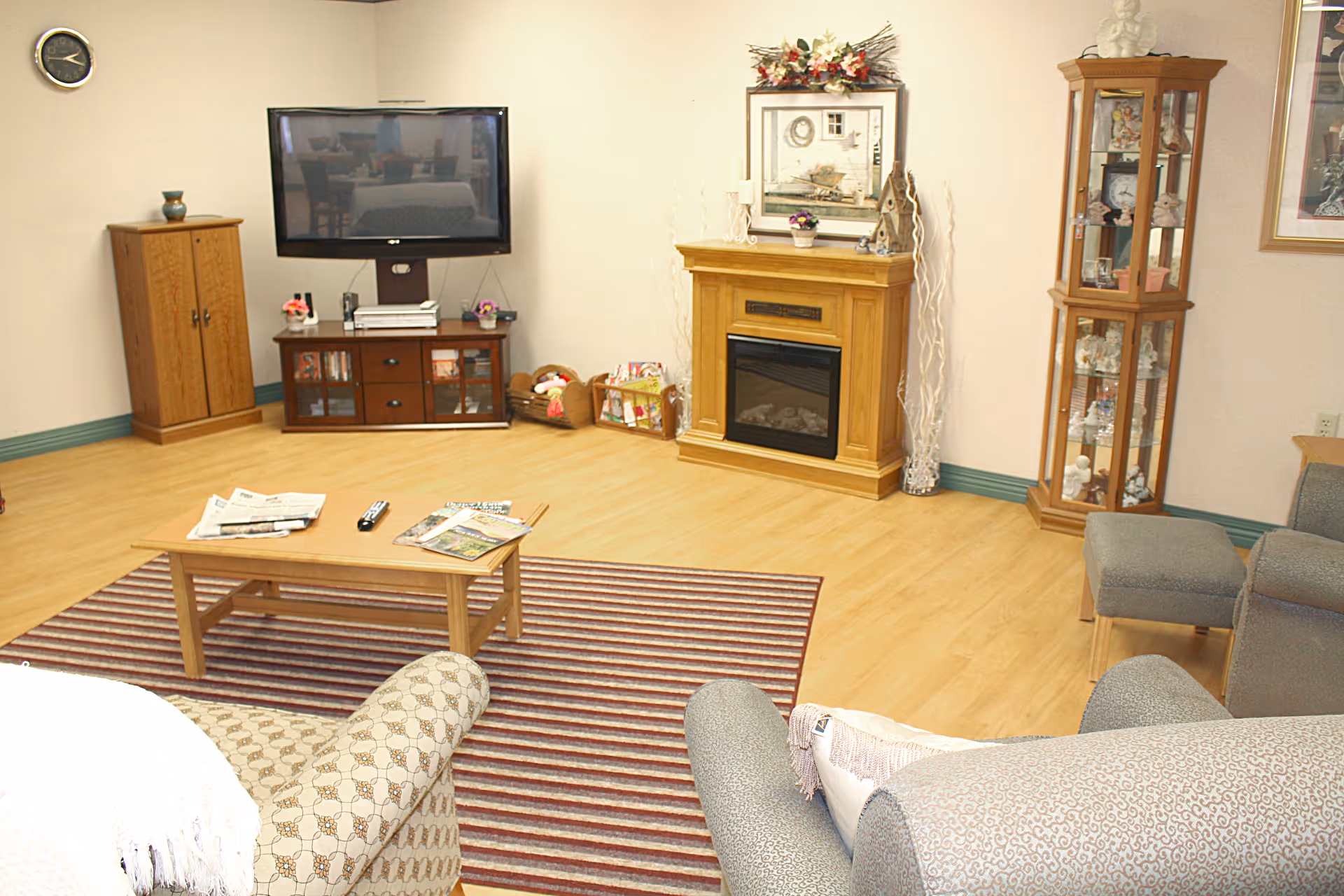 A cozy living room with a wooden floor and a striped area rug. The room features a flat-screen TV mounted on the wall above a wooden cabinet, a wooden coffee table with newspapers and a remote control on it, a fireplace with decorative items on the mantel, and a glass display cabinet filled with figurines. There are upholstered chairs and a sofa partially visible in the foreground.