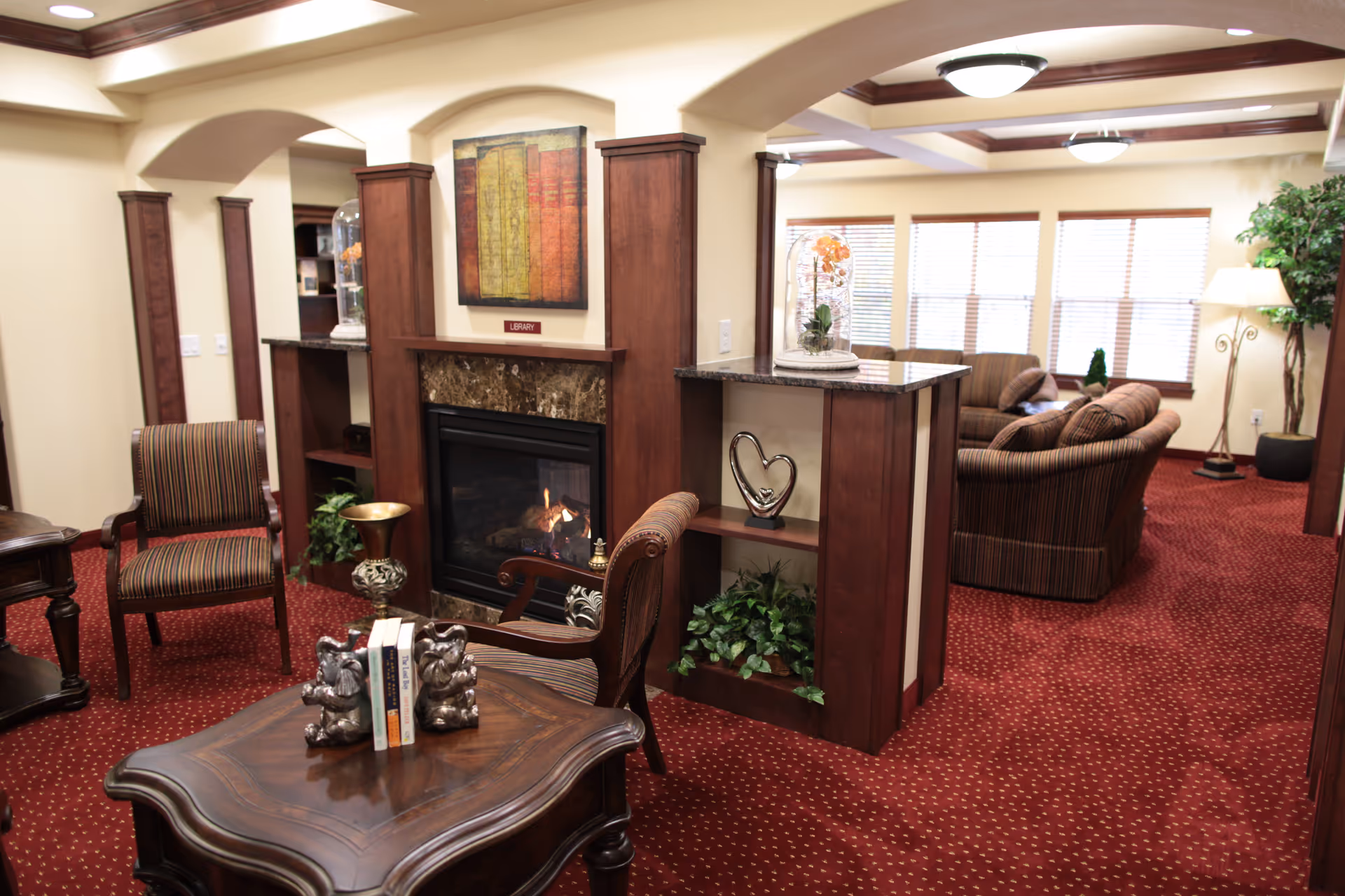 A cozy living room area in a senior living facility featuring a fireplace with a marble surround and wooden mantel. The room has red patterned carpet, wooden furniture including a coffee table with books and decorative bookends, striped upholstered chairs, and a sofa set in the background near large windows with blinds. There are decorative plants and artwork on the walls, creating a warm and inviting atmosphere.