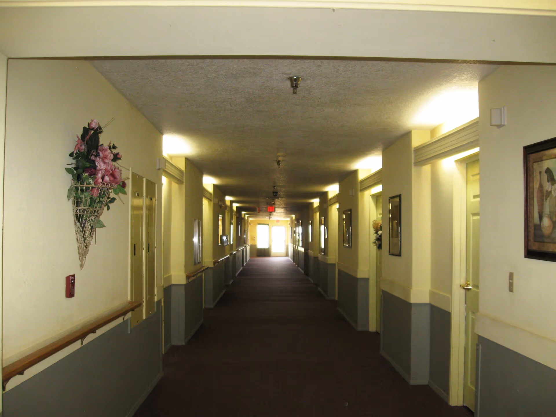 A long, well-lit hallway in an assisted living facility with beige and gray walls, handrails on both sides, framed pictures, and floral wall decorations. Doors line both sides of the corridor, and an exit door is visible at the far end.