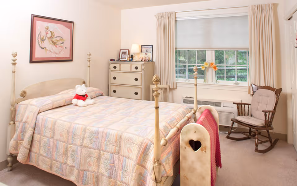 A cozy bedroom with a wooden four-poster bed covered in a pastel patchwork quilt. A white teddy bear with a red scarf sits on the bed. There is a wooden dresser with framed photos and a lamp on top, a window with beige curtains and a pleated shade, and a wooden rocking chair with a cushion near the window. A pink blanket is draped over the footboard of the bed.