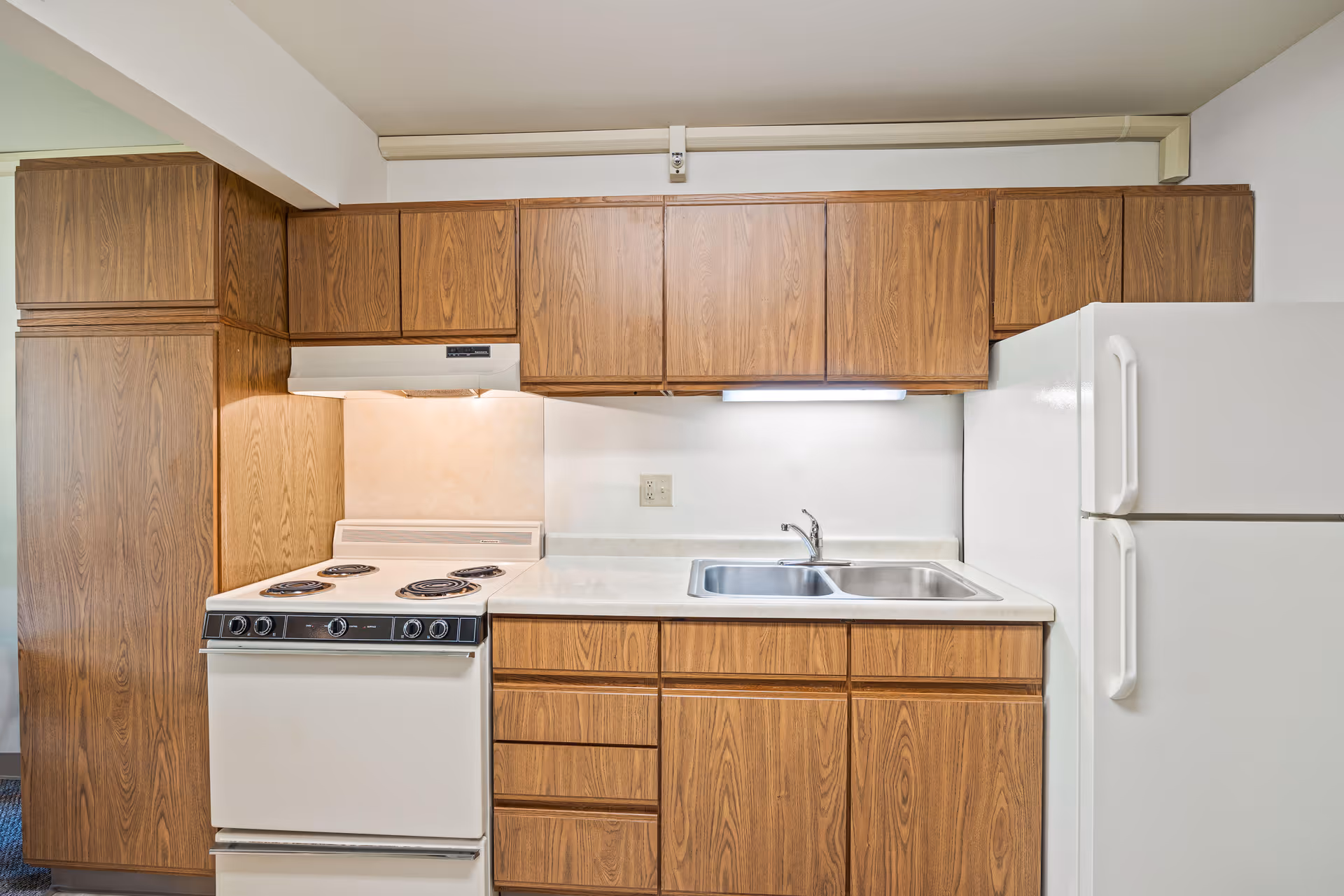 Compact kitchen with wood cabinets, an electric stove, a double sink, and a white refrigerator.