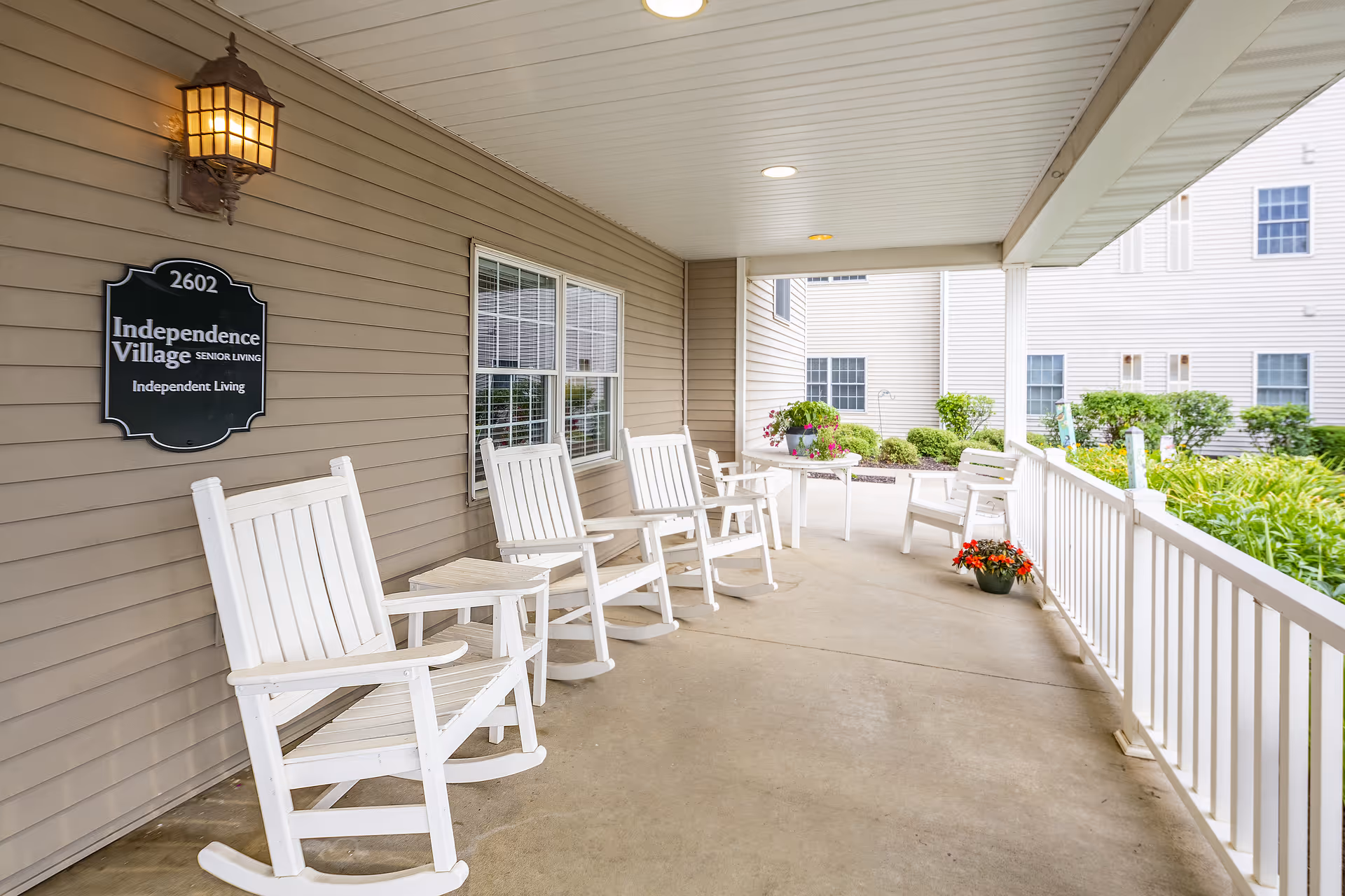 Covered outdoor porch area at Independence Village of Pella with white rocking chairs and small tables along the wall, a hanging lantern light, potted plants, and a view of the garden and building exterior.