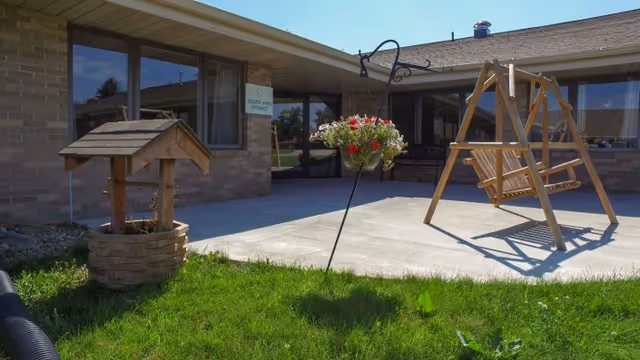A sunny courtyard with a wooden porch swing, hanging flower basket, and a decorative wishing-well planter in front of a low brick building.