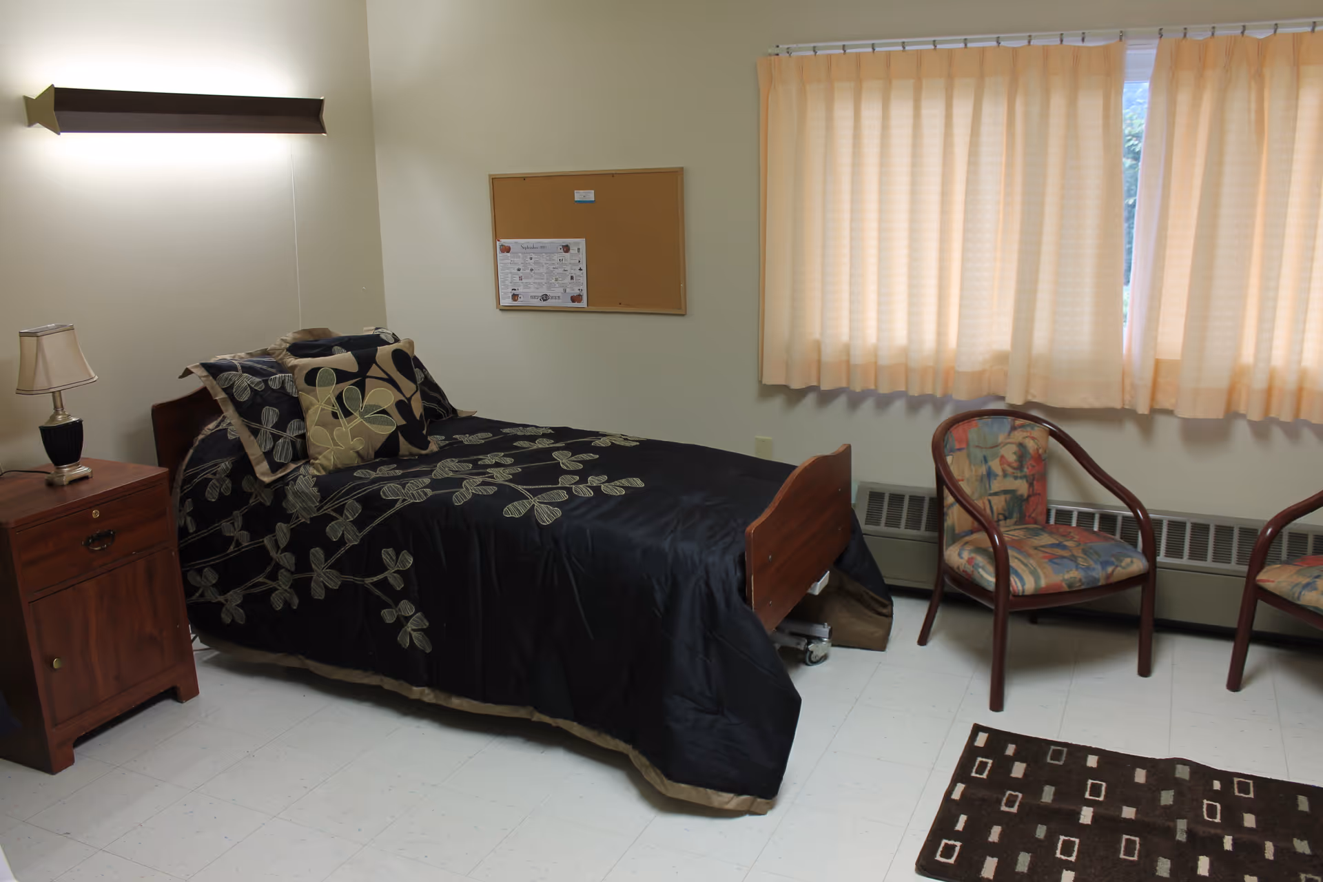 A simple bedroom in a healthcare facility with a single bed covered in a black bedspread with floral patterns, a wooden nightstand with a lamp, two patterned chairs near a window with beige curtains, a cork bulletin board on the wall, and a small patterned rug on the floor.