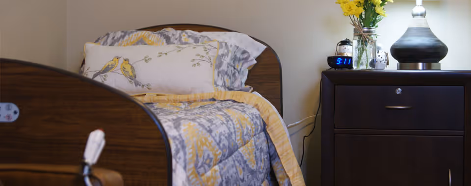 A neatly made bed with a wooden headboard and a comforter featuring a yellow and gray pattern. There is a pillow with a bird design on it. Next to the bed is a dark wooden nightstand with a digital clock showing 9:11, a vase with yellow flowers, a small snowman decoration, and a lamp with a round base.