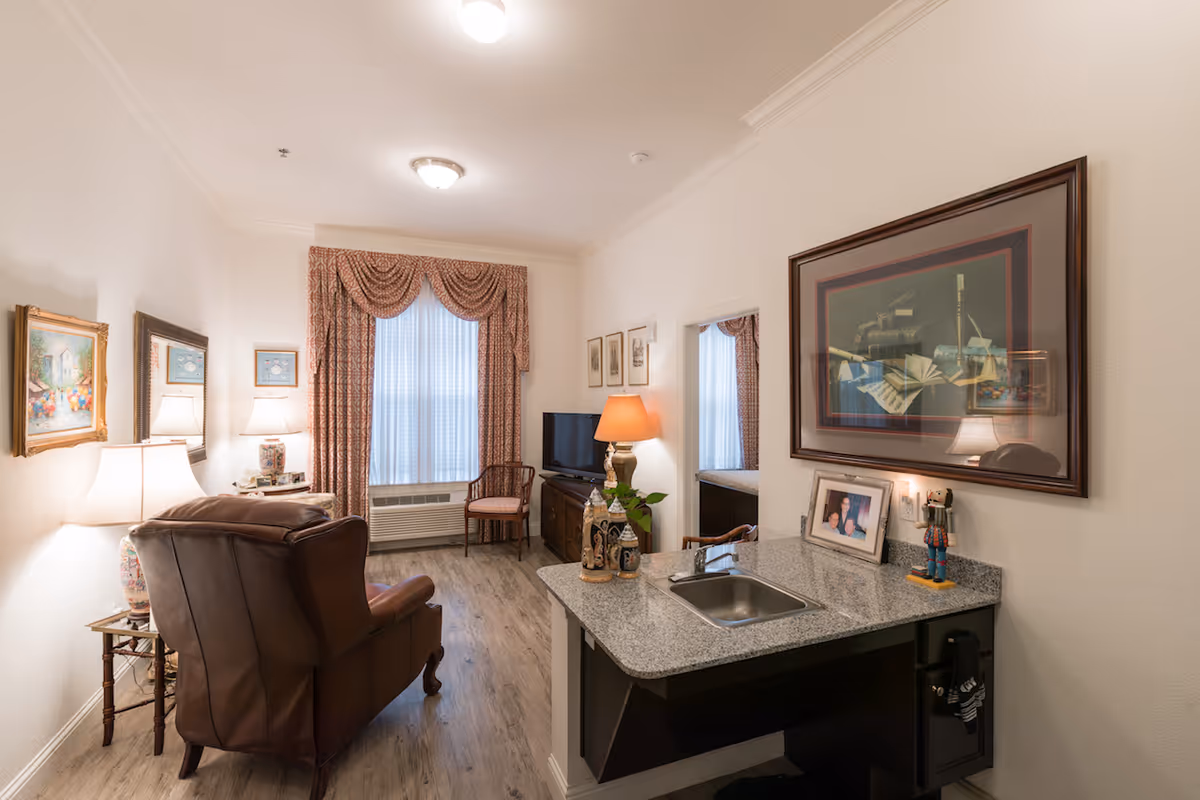 A cozy living room area in a senior living facility featuring a brown leather armchair, a wooden chair near a window with patterned curtains, a TV on a wooden stand, and a small granite countertop with a sink. The room is decorated with framed artwork, table lamps, and decorative items, with light wood flooring and white walls.