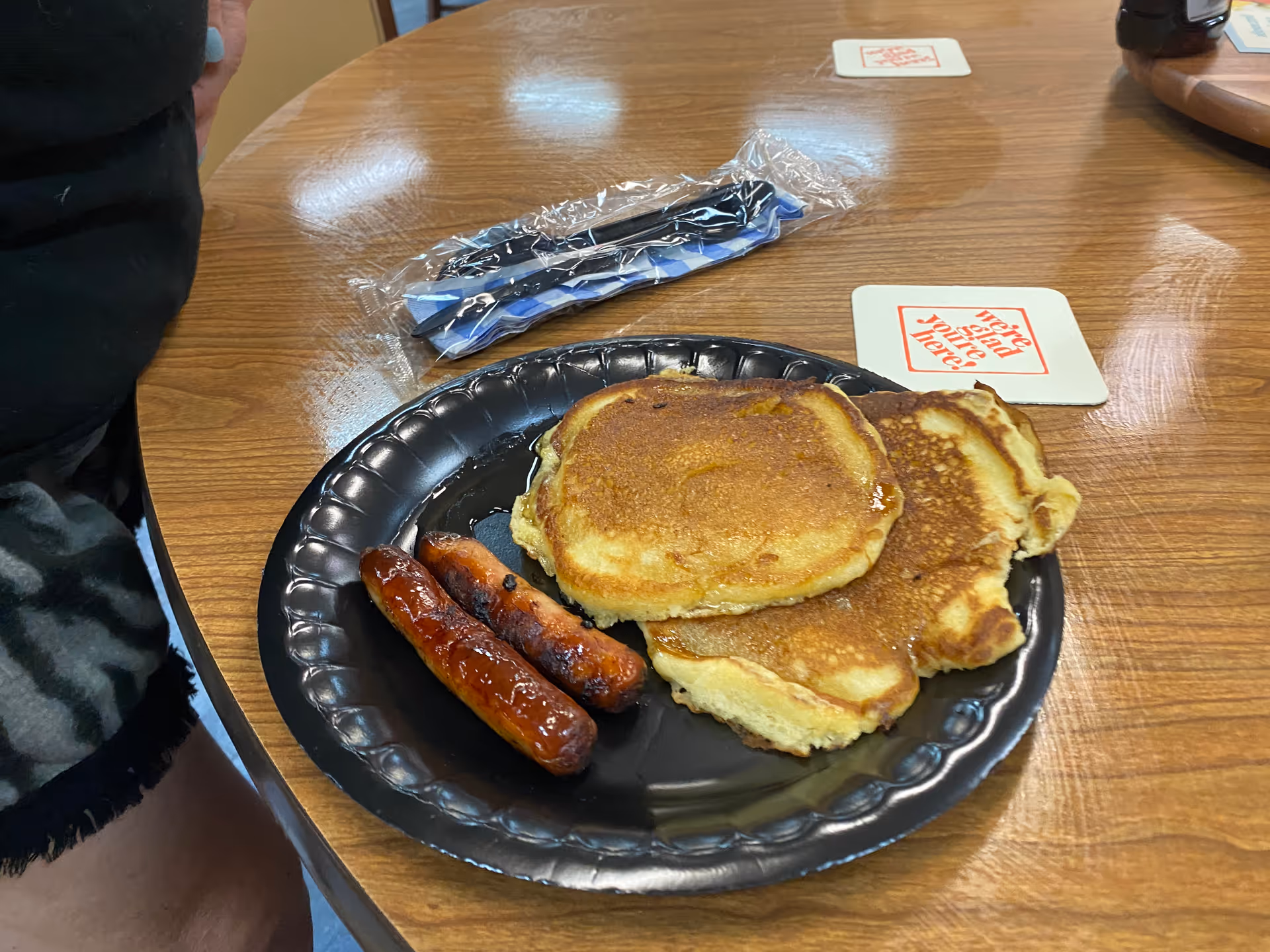 A black disposable plate on a wooden table with two pancakes and two sausage links. There is a wrapped plastic utensil set and a coaster with red text on the table nearby.