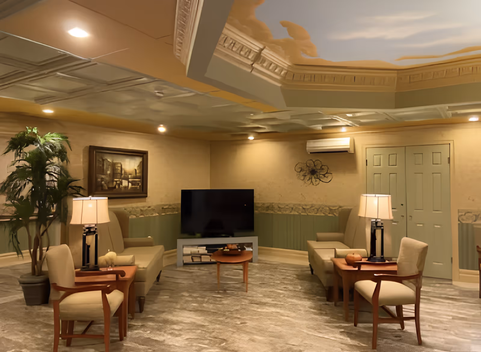 A cozy living room area with beige armchairs and wooden side tables with lamps. A flat-screen TV is placed on a stand against the wall. The room has a decorative ceiling with crown molding, a potted plant in the corner, a framed painting on the wall, and a wall-mounted air conditioning unit above a set of double doors.