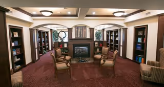 A cozy living room area with a central fireplace flanked by built-in bookshelves filled with books and decorative items. Four wooden chairs with cushions are arranged around the fireplace on a carpeted floor. The room has warm lighting and arched openings above the bookshelves.