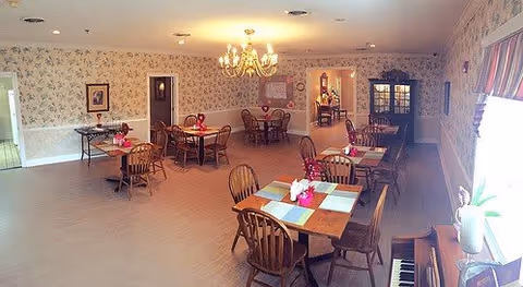 A dining room with several wooden tables and chairs arranged neatly. Each table has placemats and a small centerpiece. The room features floral wallpaper, a chandelier hanging from the ceiling, framed pictures on the walls, and a piano near a window with striped curtains. There is a doorway leading to another room in the background.