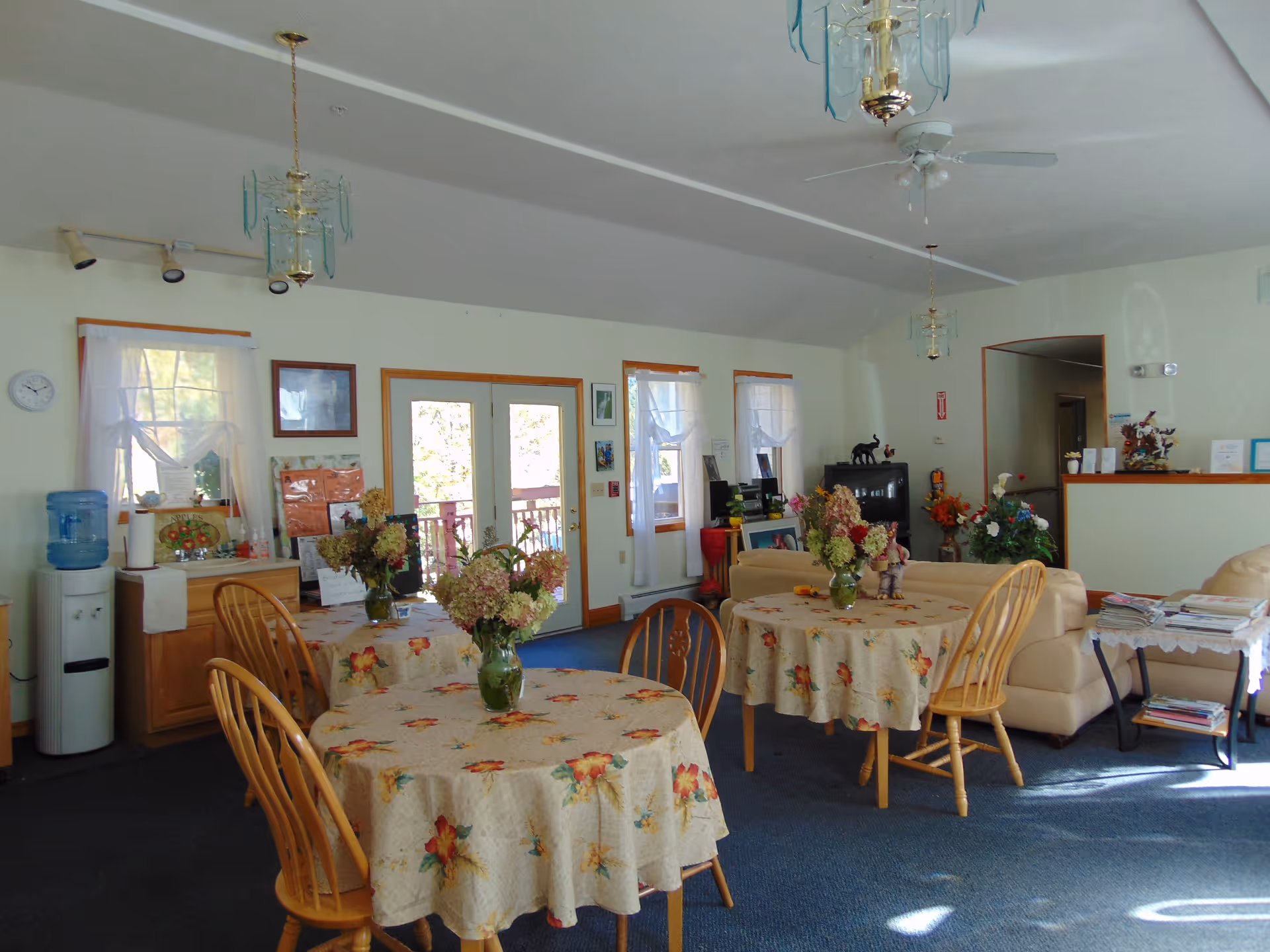 A cozy common area in an assisted living facility with round tables covered in floral tablecloths and vases of flowers. There are wooden chairs around the tables, beige sofas, a TV, and a water cooler near windows with sheer white curtains. The room has blue carpet and light-colored walls with framed pictures and decorations.