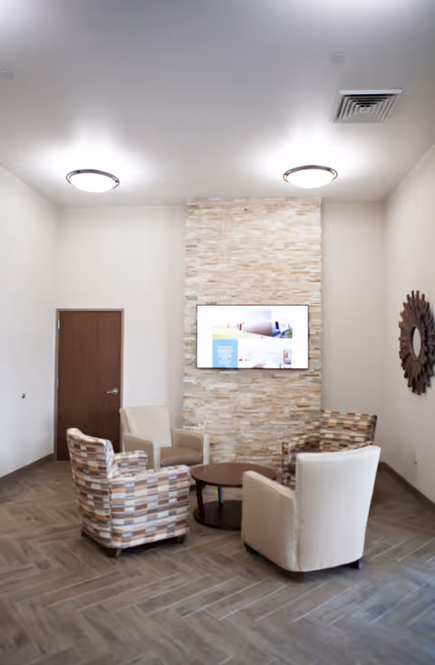 A small seating area with four armchairs arranged around a round wooden coffee table in front of a stone accent wall with a mounted flat-screen TV. The room has a wooden door on the left, a decorative round mirror on the right wall, and two ceiling lights.