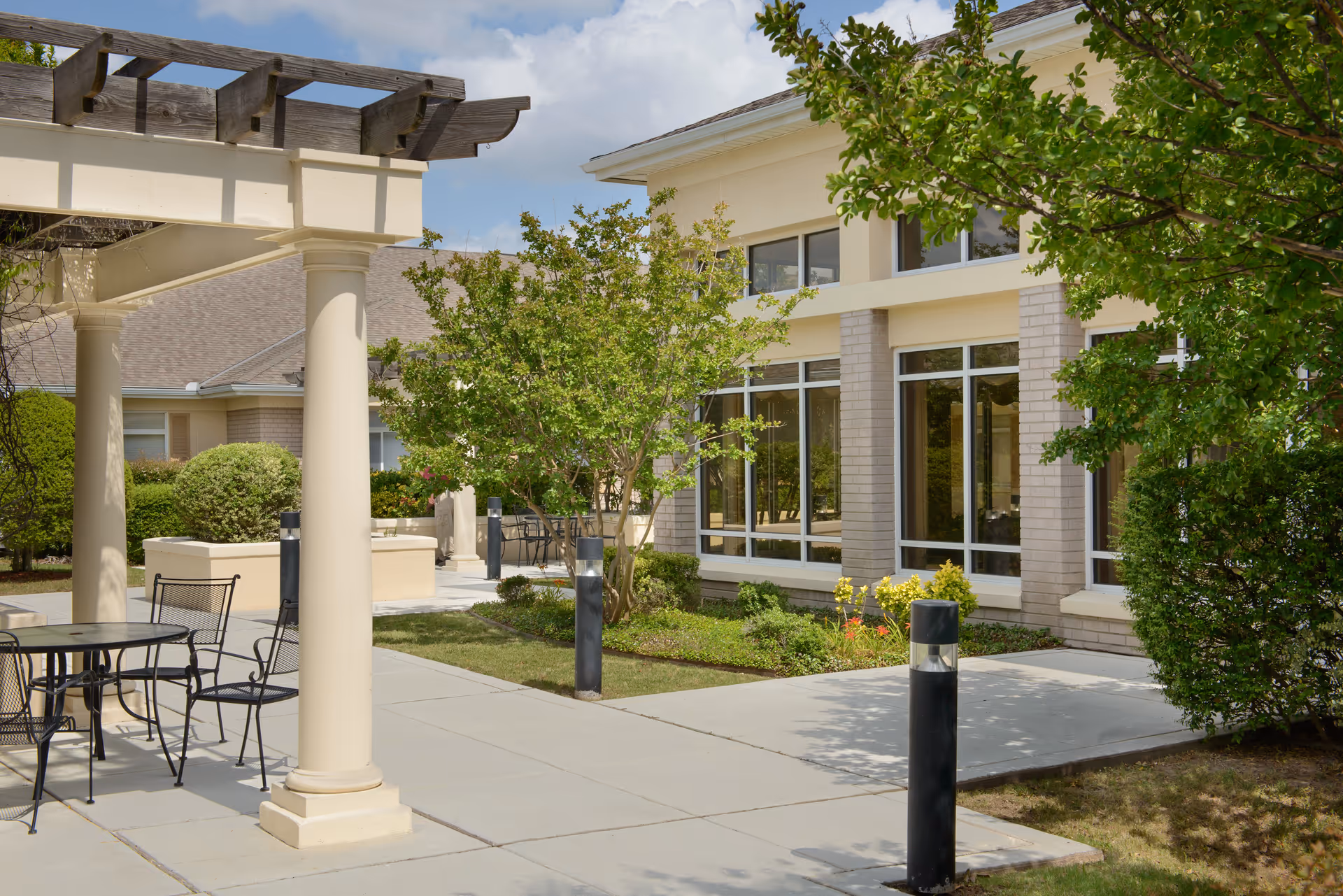 Outdoor patio area at Garden Terrace at Fort Worth featuring a pergola with columns, metal chairs and tables, a concrete walkway, landscaped bushes and trees, and a building with large windows in the background under a partly cloudy sky.