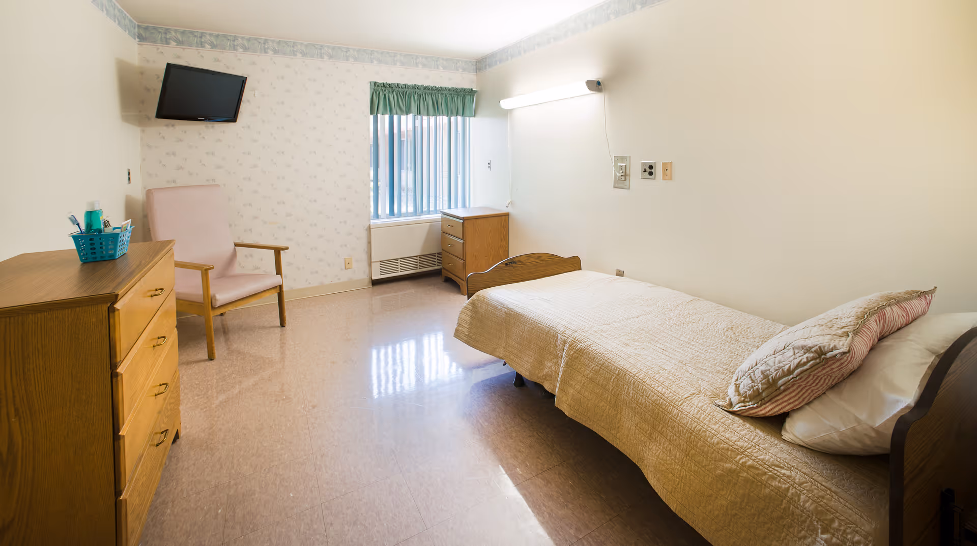 A simple nursing facility bedroom with a single bed covered with a beige quilt and pillows, a wooden dresser with a basket of toiletries, a pink cushioned chair, a small wooden chest of drawers near a window with vertical blinds, and a wall-mounted TV in the corner.