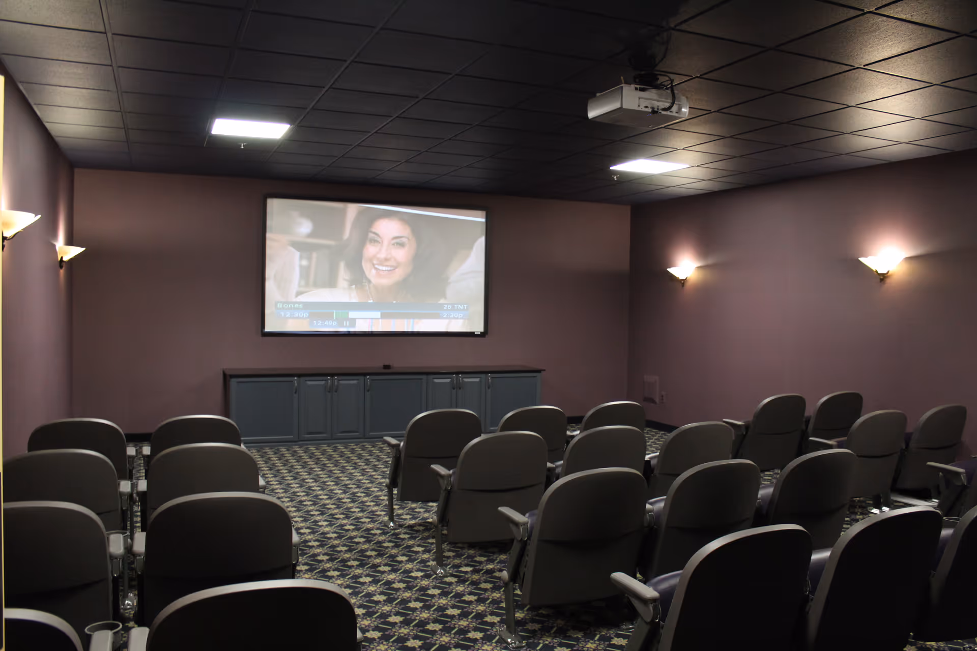 A small theater room with multiple rows of gray cushioned chairs facing a large screen on the front wall. The screen displays a woman smiling. The room has purple walls, patterned carpet, wall-mounted lights, and a ceiling-mounted projector.