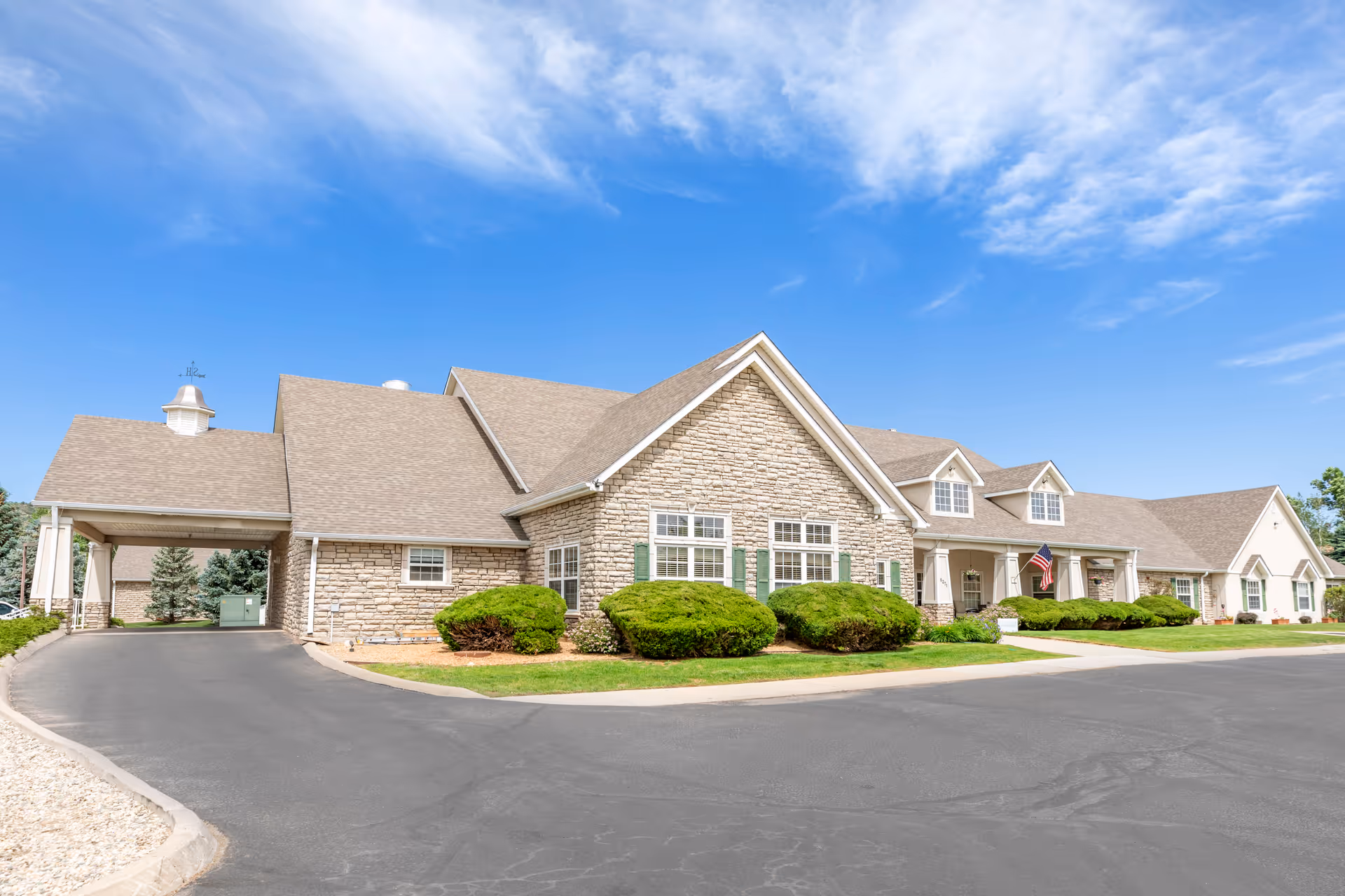 Exterior view of a single-story senior living facility building with stone and siding facade, multiple windows with green shutters, a covered entrance, well-maintained shrubs, and a clear blue sky.