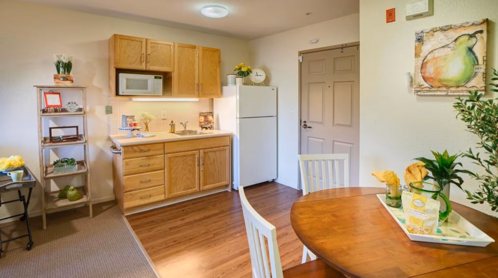 A small kitchen area with wooden cabinets, a white microwave, a white refrigerator, and a sink. There is a wooden dining table with white chairs in the foreground, decorated with a tray holding a green pitcher, glasses, and napkins. To the left, there is a wooden shelving unit with decorative items and plants. The floor transitions from carpet to wood, and a door is visible in the background with a pear-themed painting on the wall.