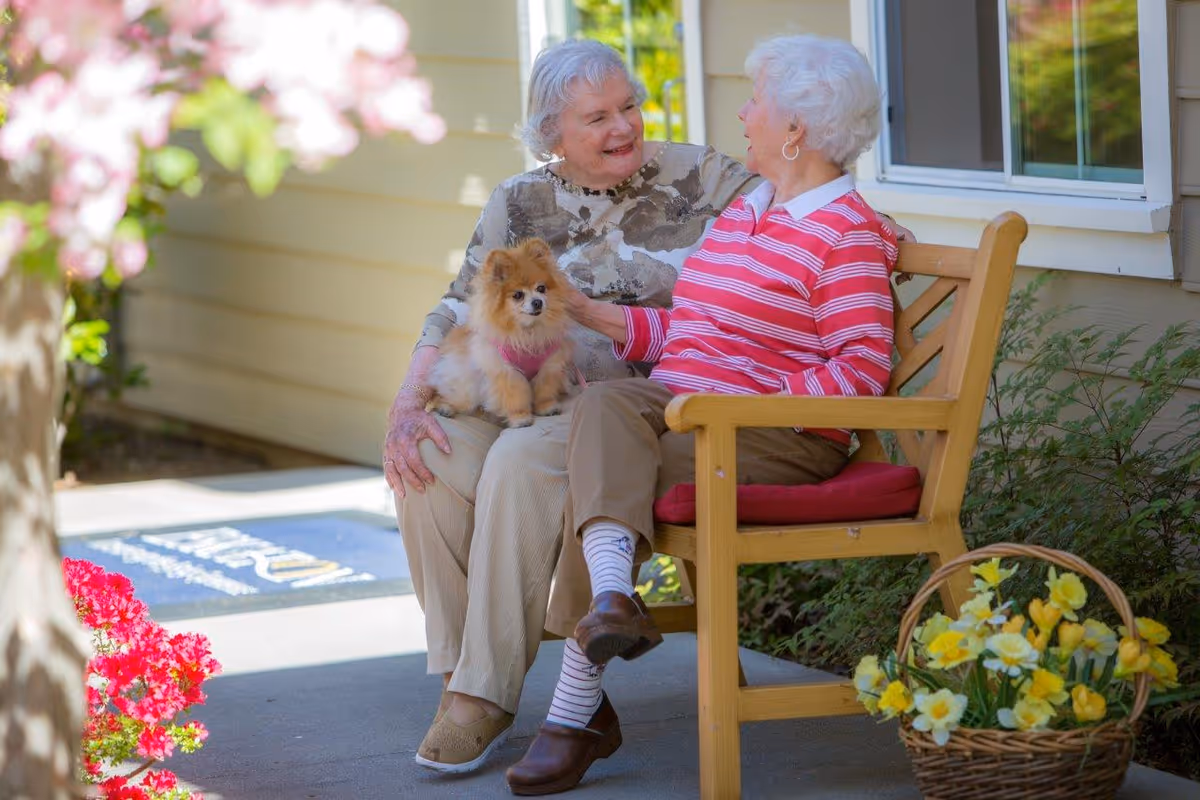 Two elderly women sitting on a wooden bench outside a building, one holding a small fluffy dog. They are smiling and engaged in conversation. There are colorful flowers in the foreground and a basket of yellow flowers on the ground nearby.