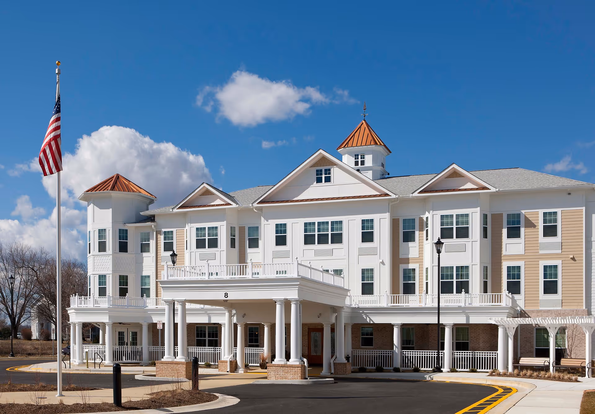 Exterior view of a senior living facility building with white and beige siding, multiple windows, and a covered entrance supported by white columns. The building has a distinctive roof with copper-colored accents and a small tower. An American flag is flying on a flagpole in front of the building under a blue sky with some clouds.