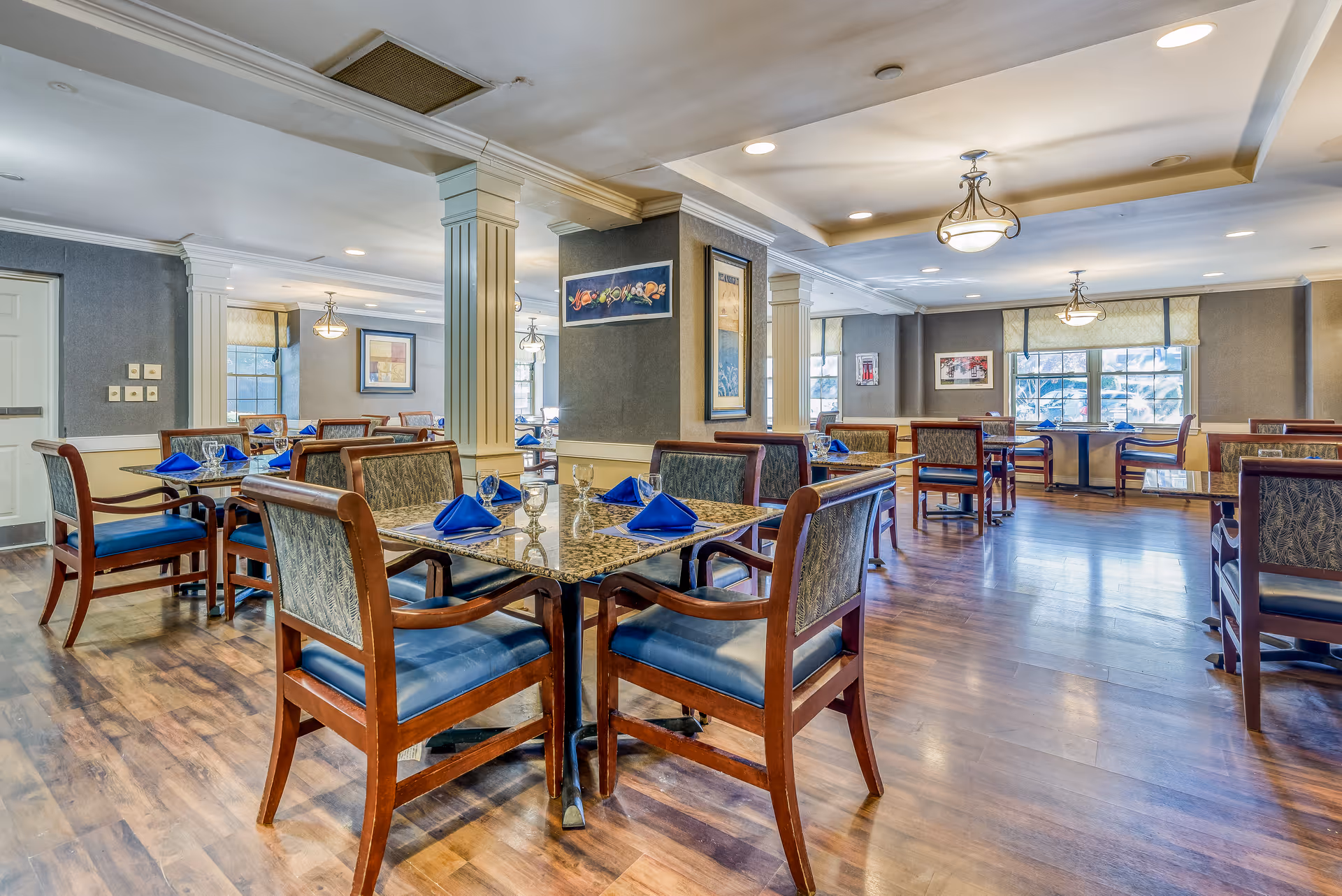 A spacious dining room with multiple tables and chairs arranged neatly. Each table has a granite top and is set with blue folded napkins and clear drinking glasses. The room features wooden flooring, decorative columns, framed artwork on the walls, and large windows allowing natural light to enter.