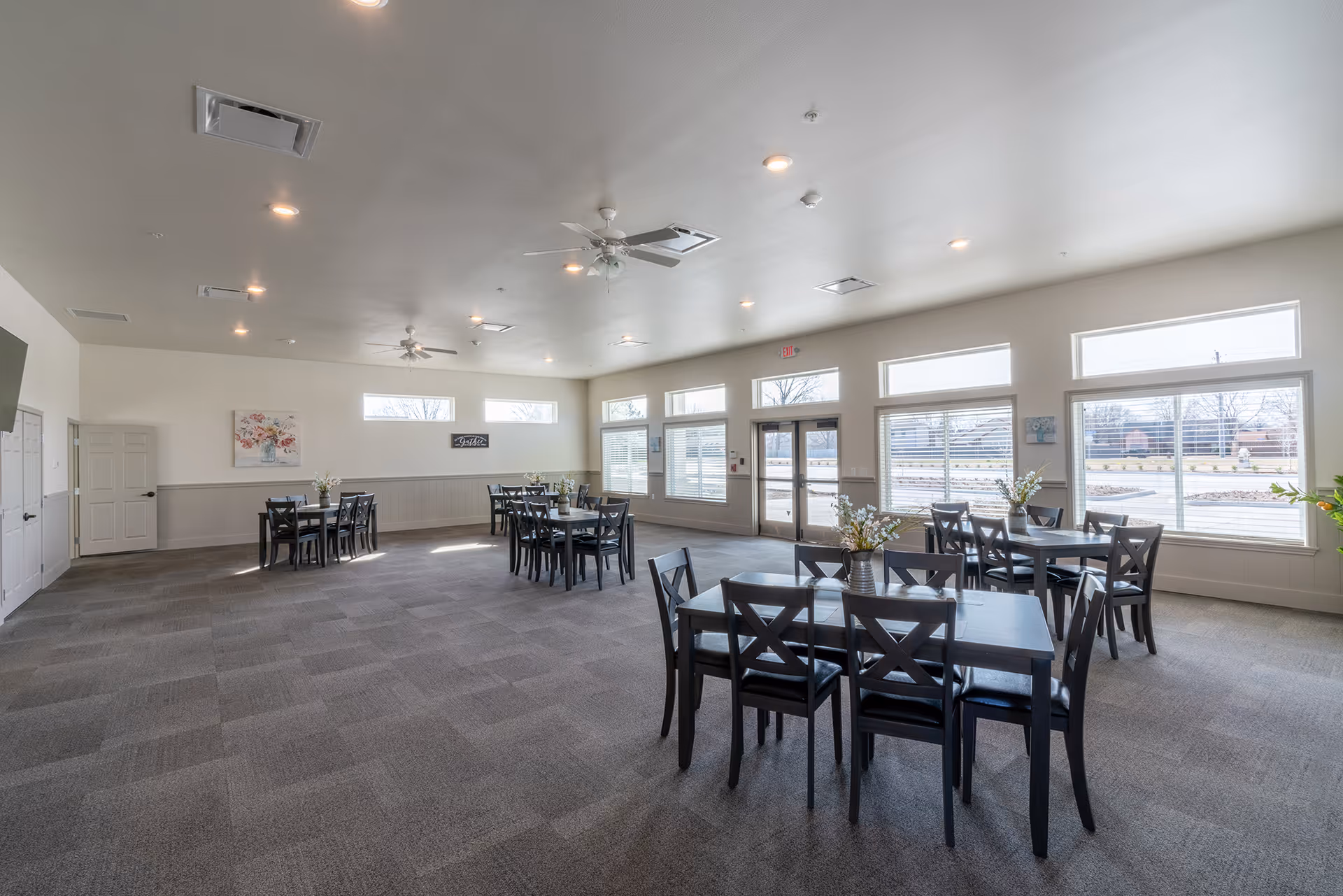 A spacious dining room with multiple black tables and chairs arranged neatly. Each table has a small vase with flowers. The room has large windows allowing natural light to fill the space, light-colored walls, and ceiling fans.
