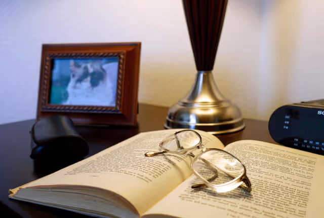 Reading glasses rest on an open book on a bedside table next to a lamp, framed photo, and alarm clock.
