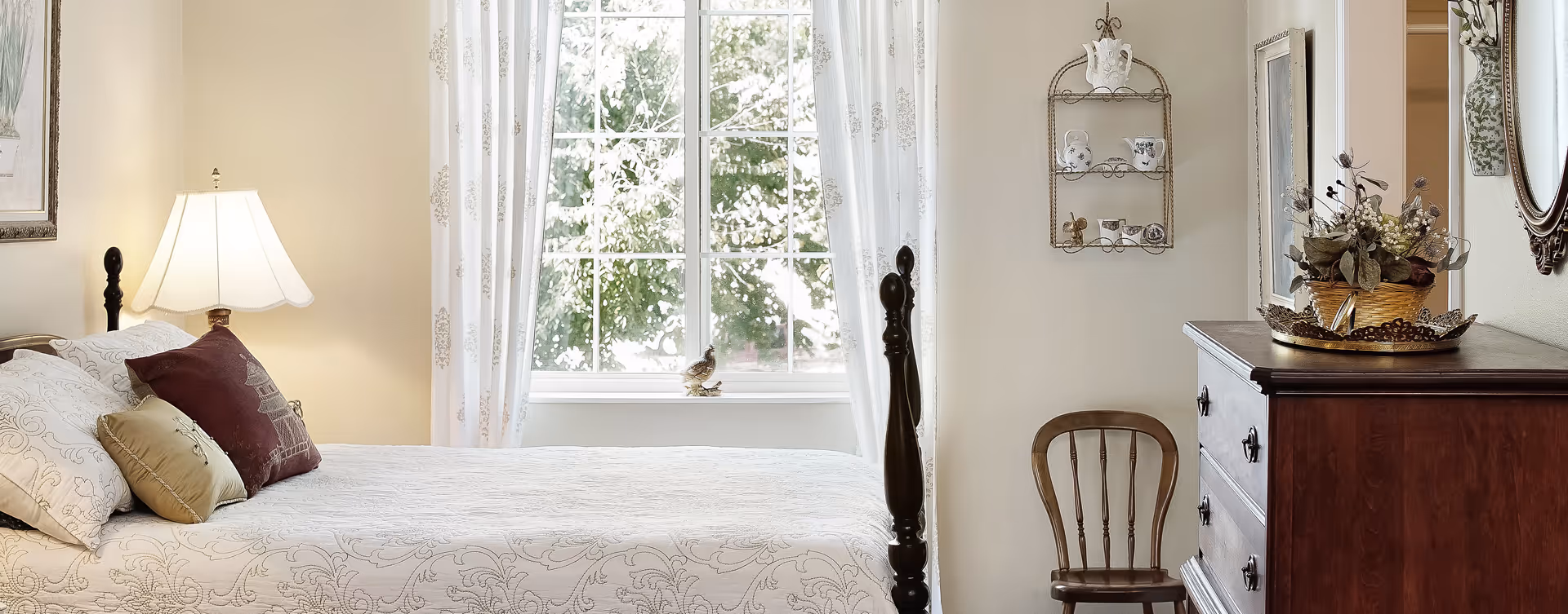 Sunlit traditional bedroom featuring a bed with decorative pillows, a bedside lamp, a window with sheer curtains, and a wooden dresser with a chair.