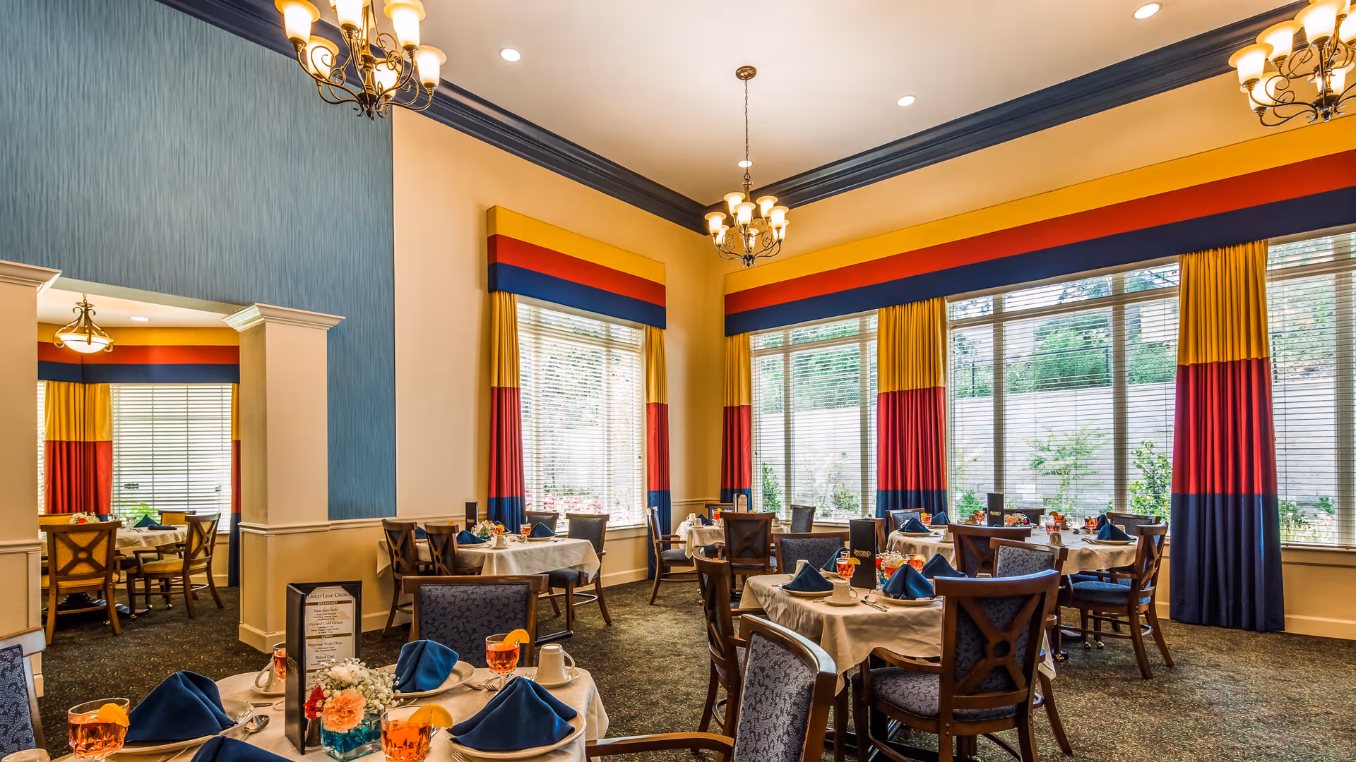 A bright dining room with multiple round tables covered with white tablecloths, each set with blue napkins, glasses with orange beverages, and floral centerpieces. The room features large windows with colorful curtains in yellow, red, and blue, and elegant chandeliers hanging from the ceiling.
