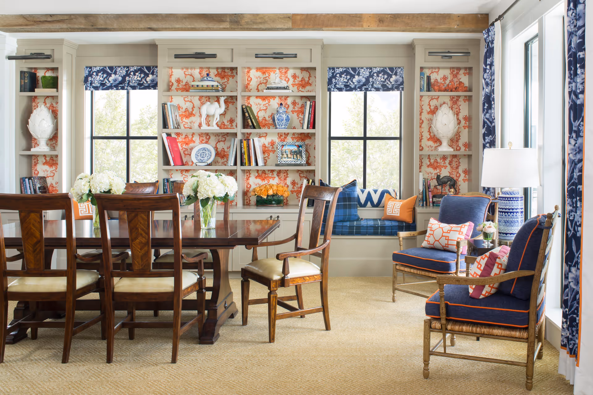 A bright and elegant living room and dining area featuring a dark wooden dining table with six chairs, two blue cushioned armchairs with patterned pillows, a window seat with blue plaid cushions and decorative pillows, built-in bookshelves with various books and decorative items, floral curtains, and a table lamp on a side table.