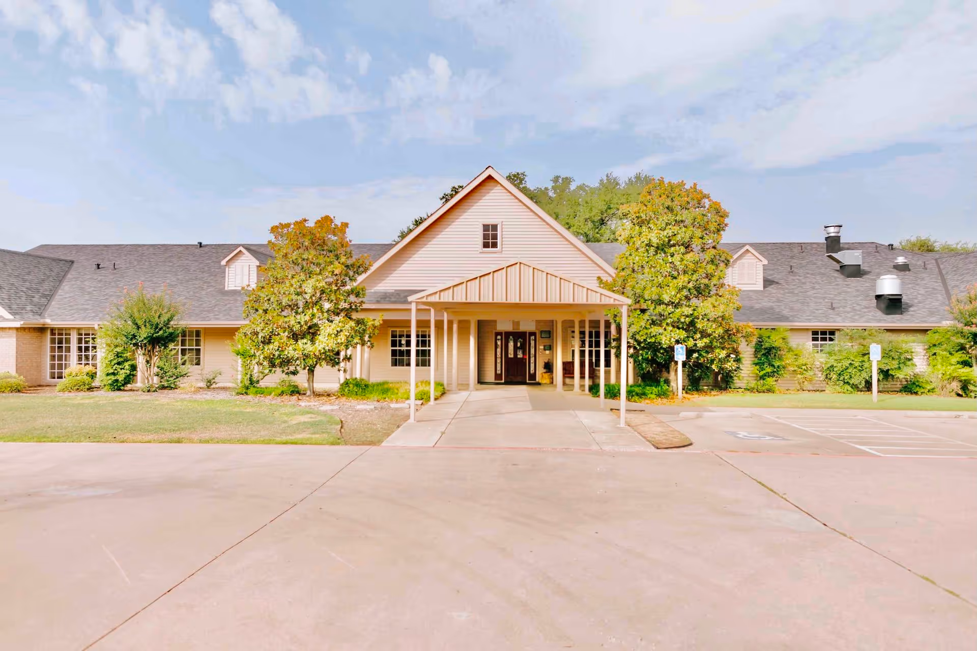 Front exterior view of a single-story senior living facility building with a covered entrance, surrounded by green trees and bushes, under a partly cloudy sky.