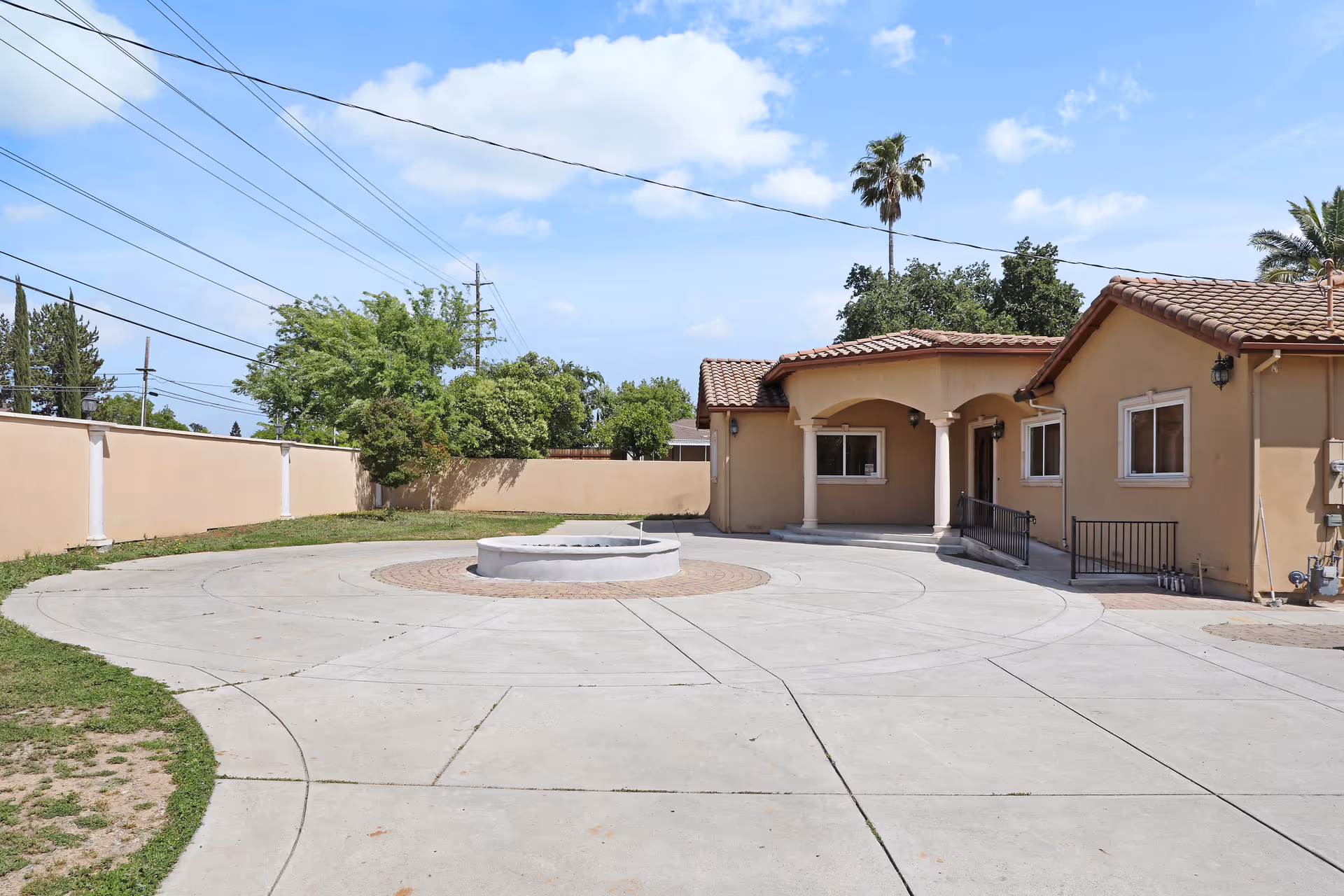 Outdoor view of a single-story beige building with a tiled roof, surrounded by a concrete circular driveway and a small round raised planter in the center. The area is enclosed by a beige wall with some greenery and trees visible behind it under a partly cloudy blue sky.