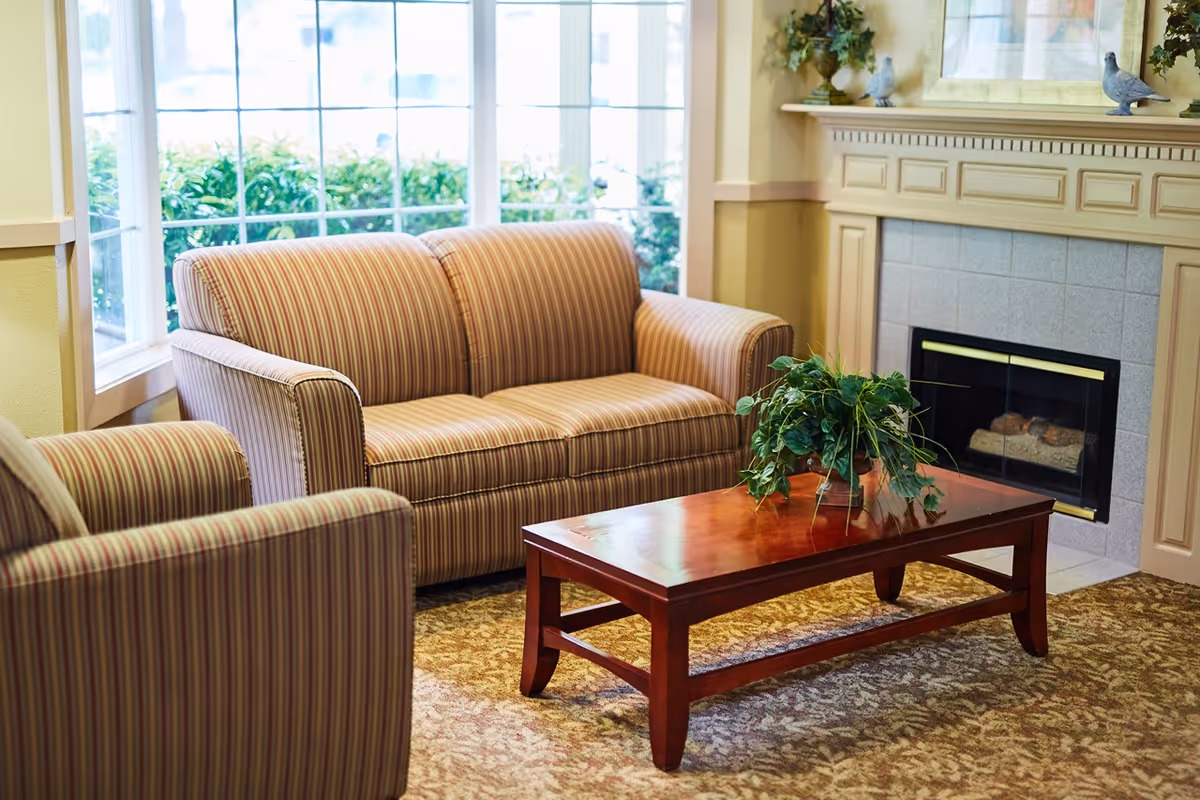 A cozy living room area with a striped sofa and armchair, a wooden coffee table with a green potted plant, a fireplace with a decorative mantel, and large windows letting in natural light.
